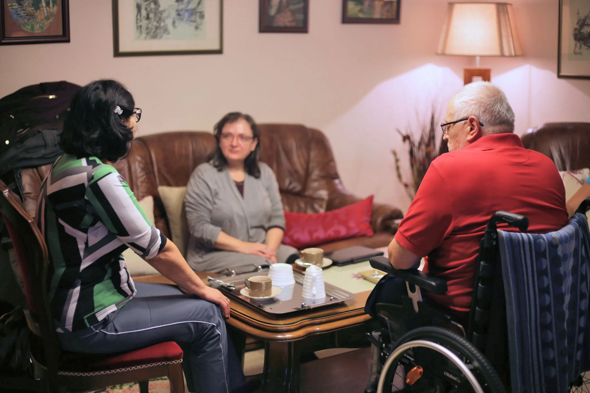 A group of four people, including a woman in a wheelchair, sitting around a coffee table engaged in conversation in a cozy living room decorated with framed pictures and a lamp.