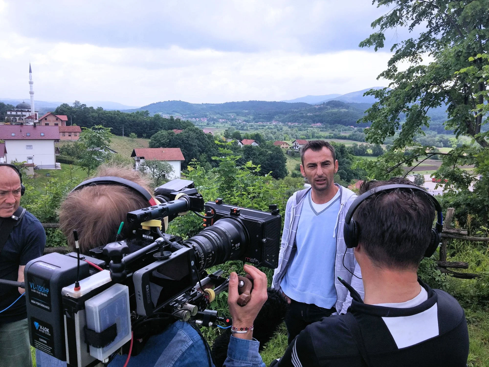 Group of people, including a man in a white shirt, standing outdoors on a hillside with a scenic view of houses, trees, and mountains in the background, filming or photographing with professional camera equipment.