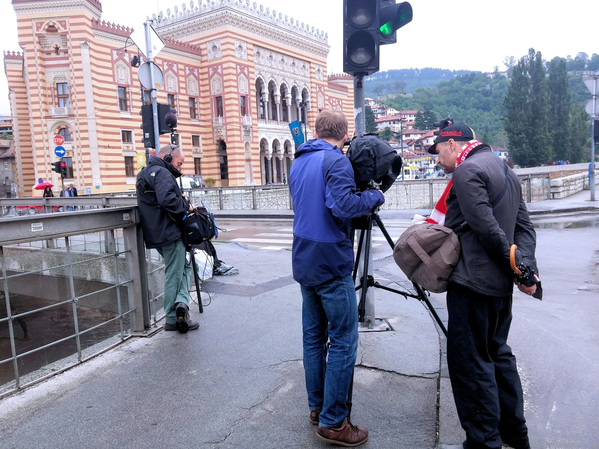 A group of four men, two with headphones, stand on a street corner in front of a historic building with intricate architectural details. They are preparing to film or record, with one man operating a camera on a tripod. Traffic lights and street sign