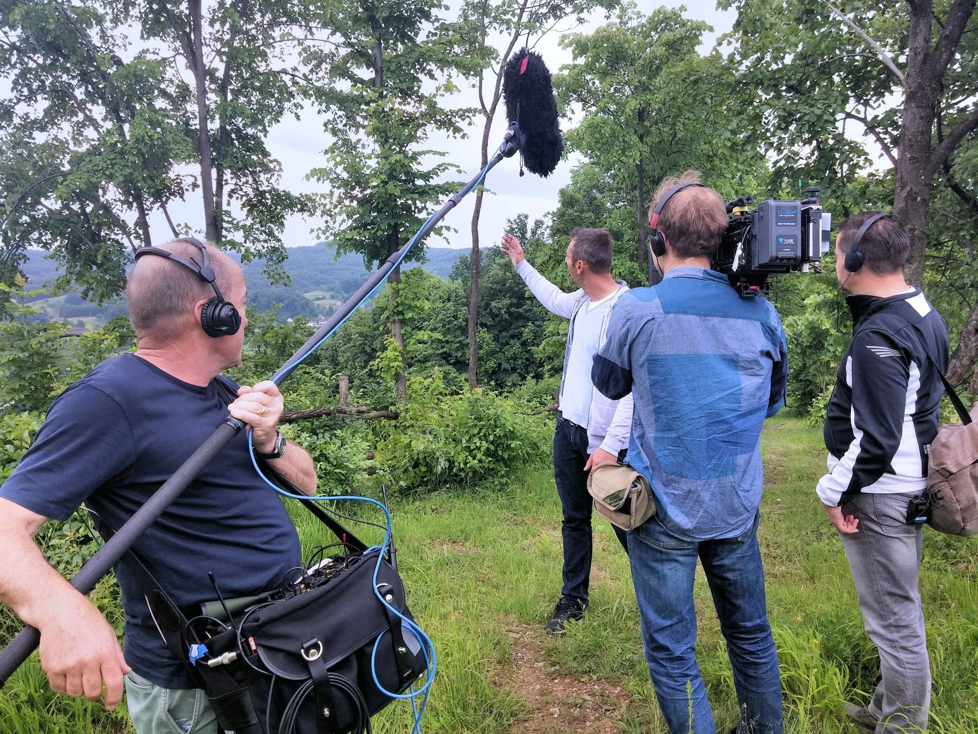 Group of people filming outdoors near trees, with a black furry creature on a stick suspended in the air.