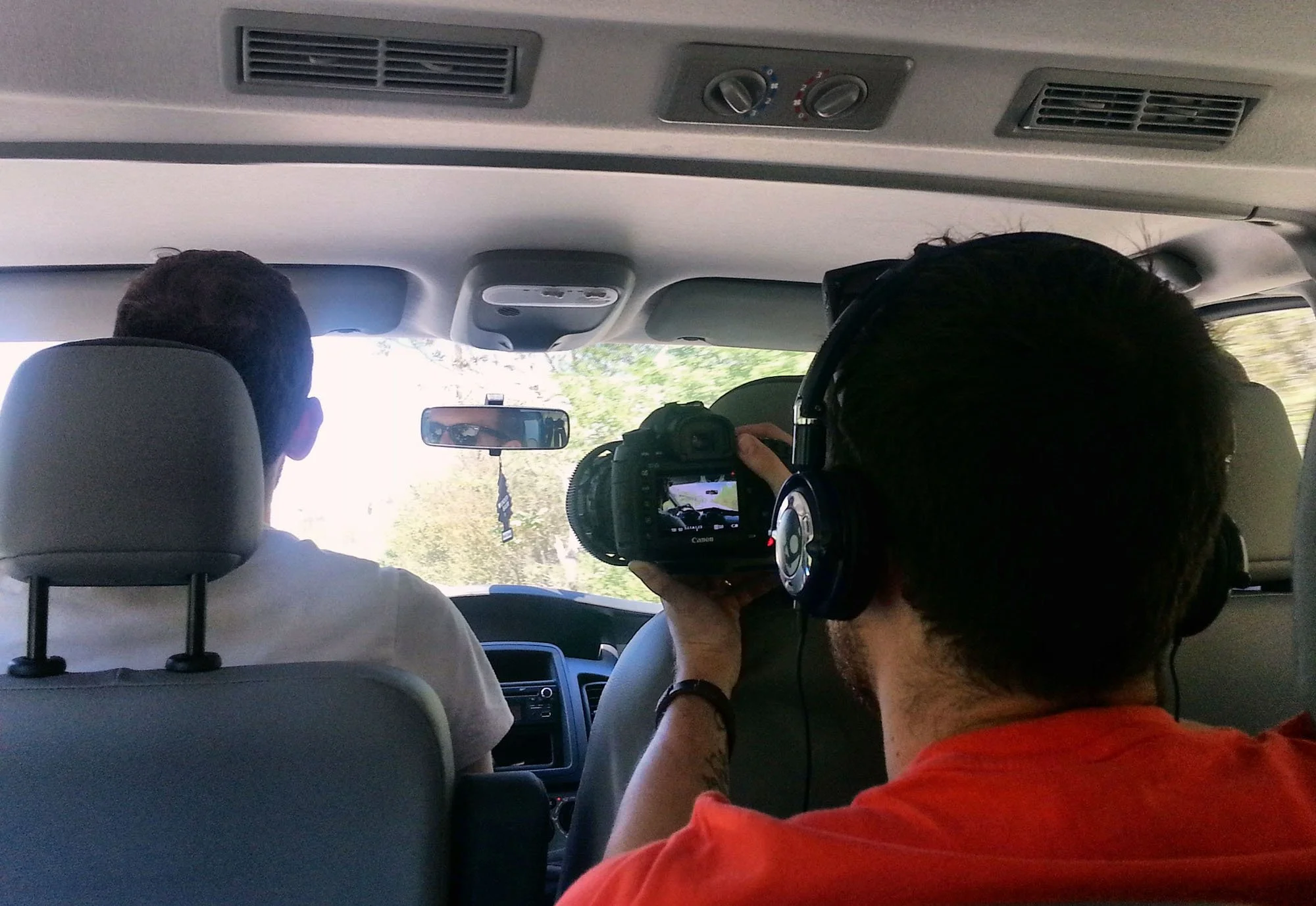 Inside a vehicle with passengers, a person is taking a photograph, and others are seated. The vehicle has ceiling air vents, air conditioning controls, and a view of a sunny day outside.