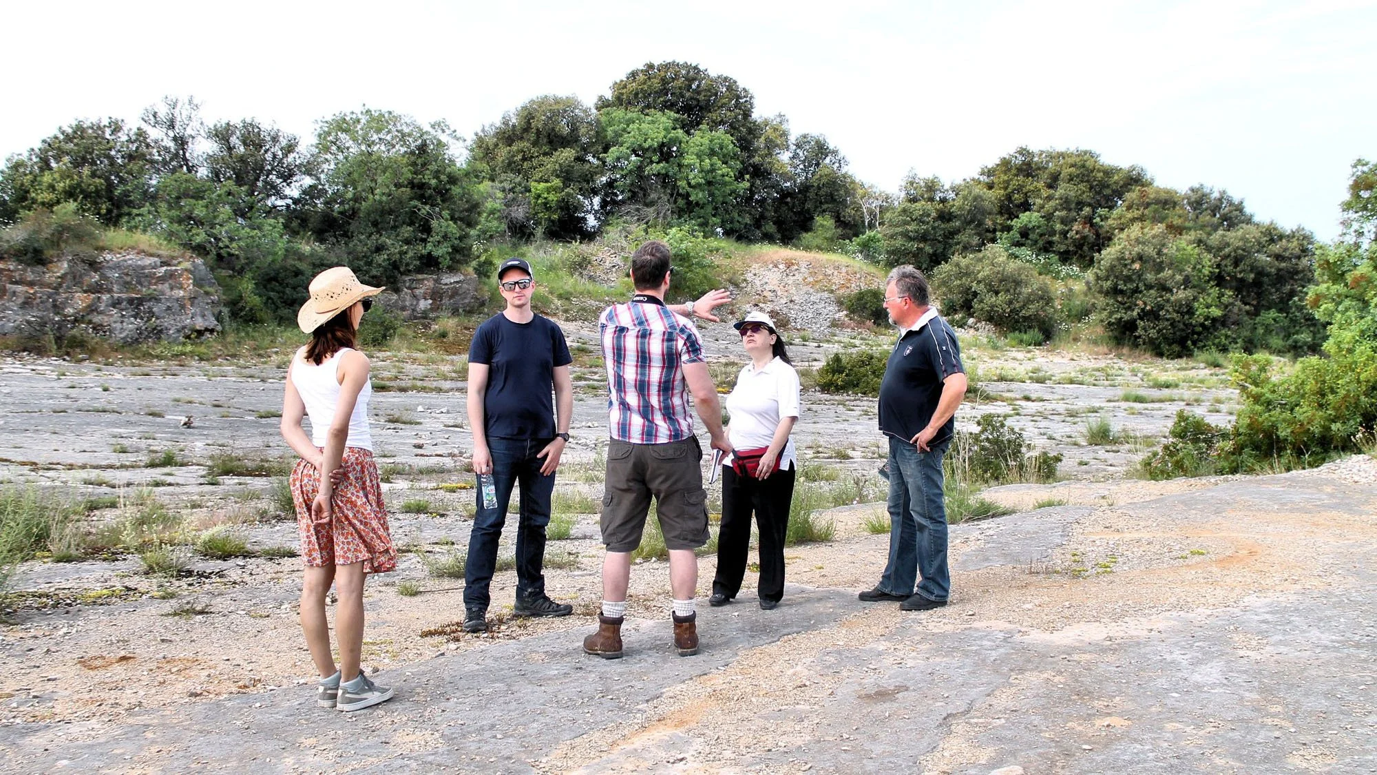 A group of six people standing outdoors on rocky terrain with trees in the background, engaged in conversation, some wearing sunglasses and hats, in casual attire.