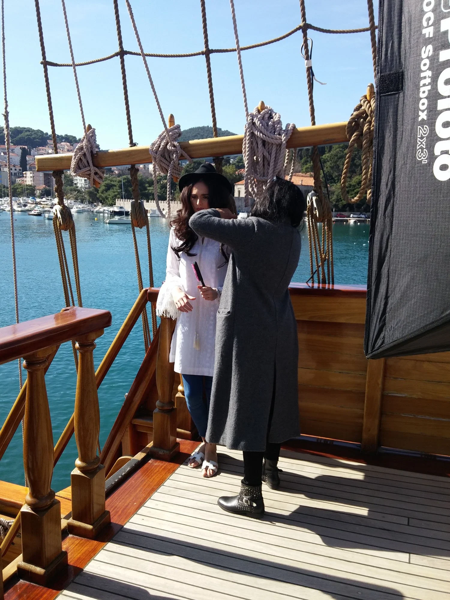 Two women on a wooden boat with rope and wood rigging, one woman in a white dress and black hat holding a hairbrush, the other woman in a gray coat and black boots facing her, with a harbor full of boats and a hillside town in the background.