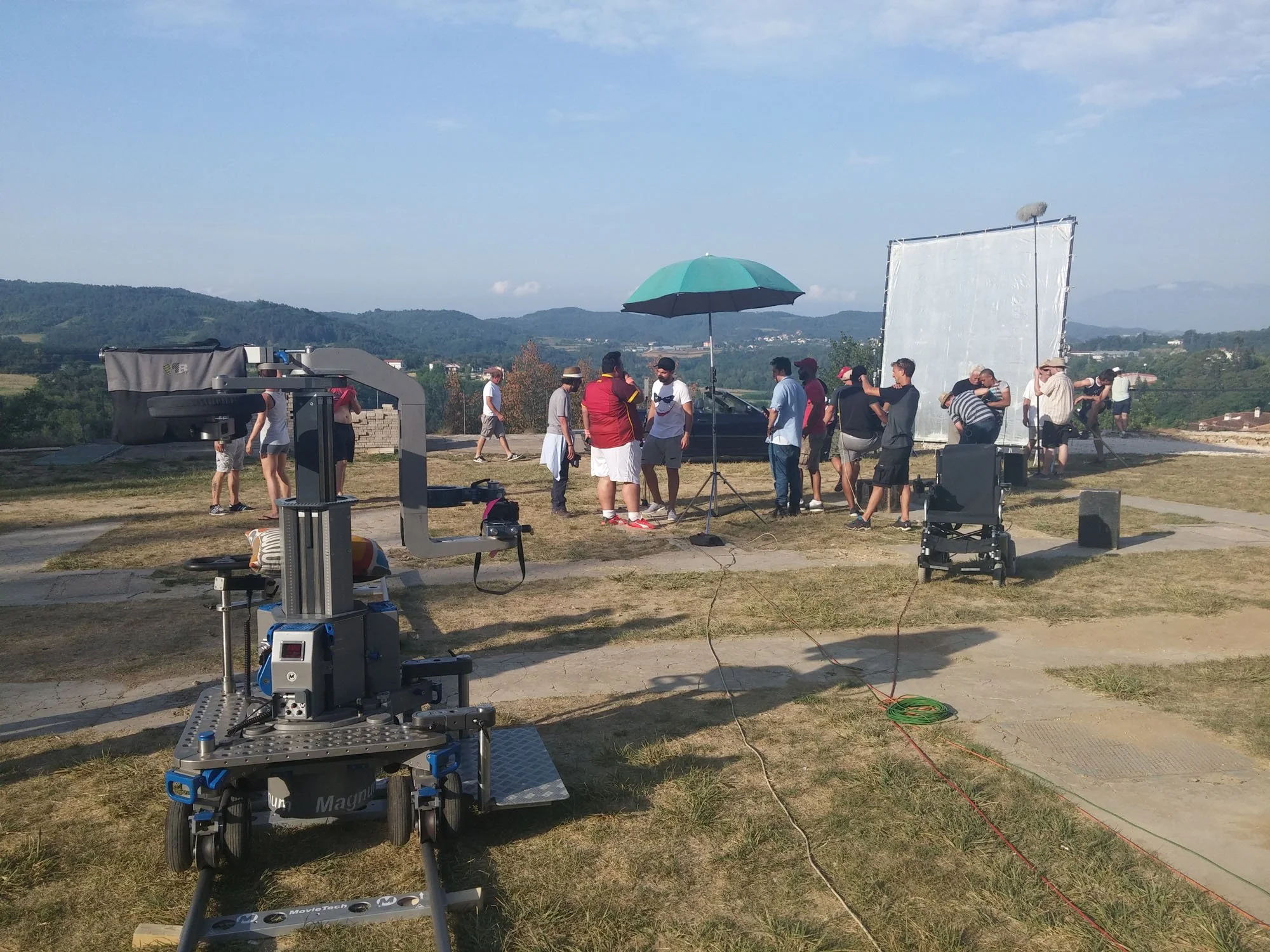 A film crew setting up equipment outdoors with mountains in the background, including a camera, lighting, and a large reflector.