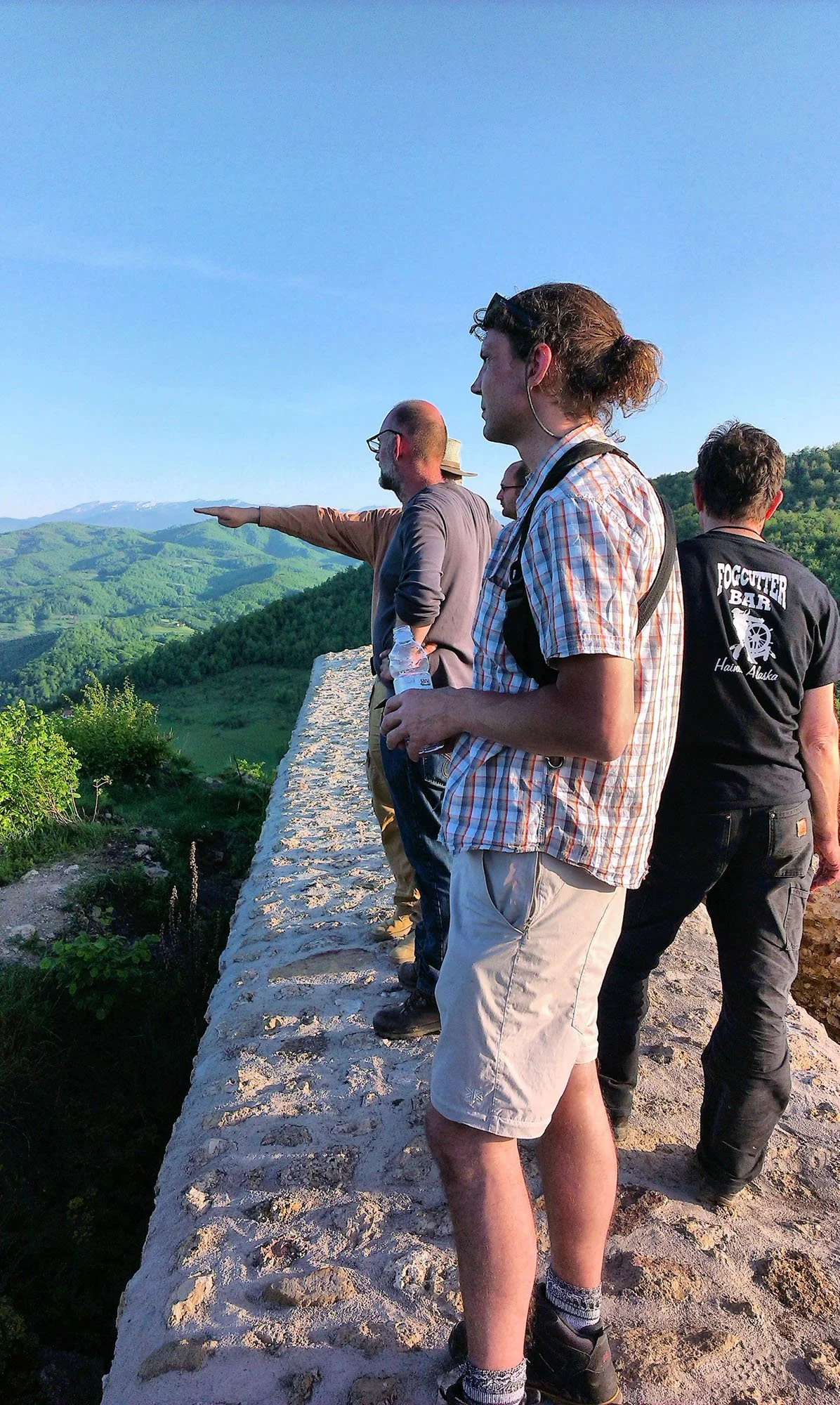 Group of people standing on a stone wall overlooking green hills, with one man pointing into the distance, in a scenic outdoor setting on a clear day.