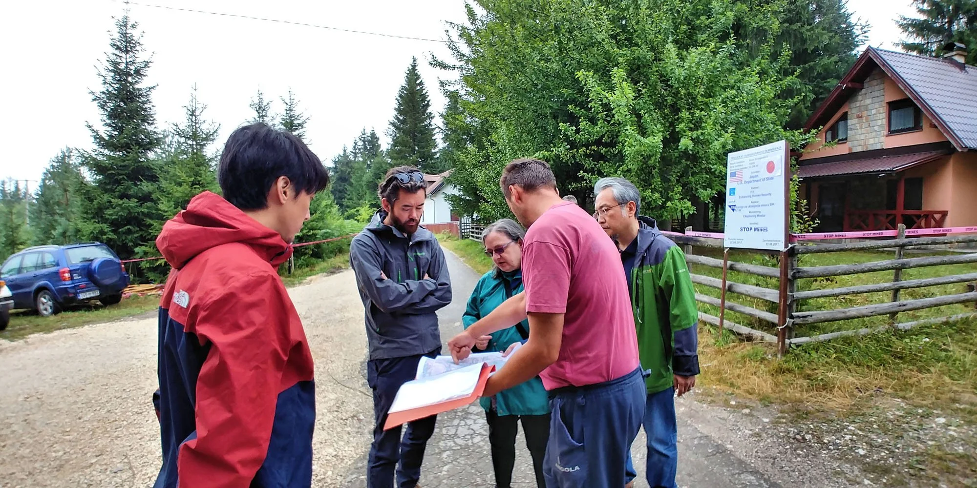 A group of people gathered outdoors reviewing documents near a wooden fence with a sign that says 'STOP MINES'. There are trees, a house, and cars in the background.