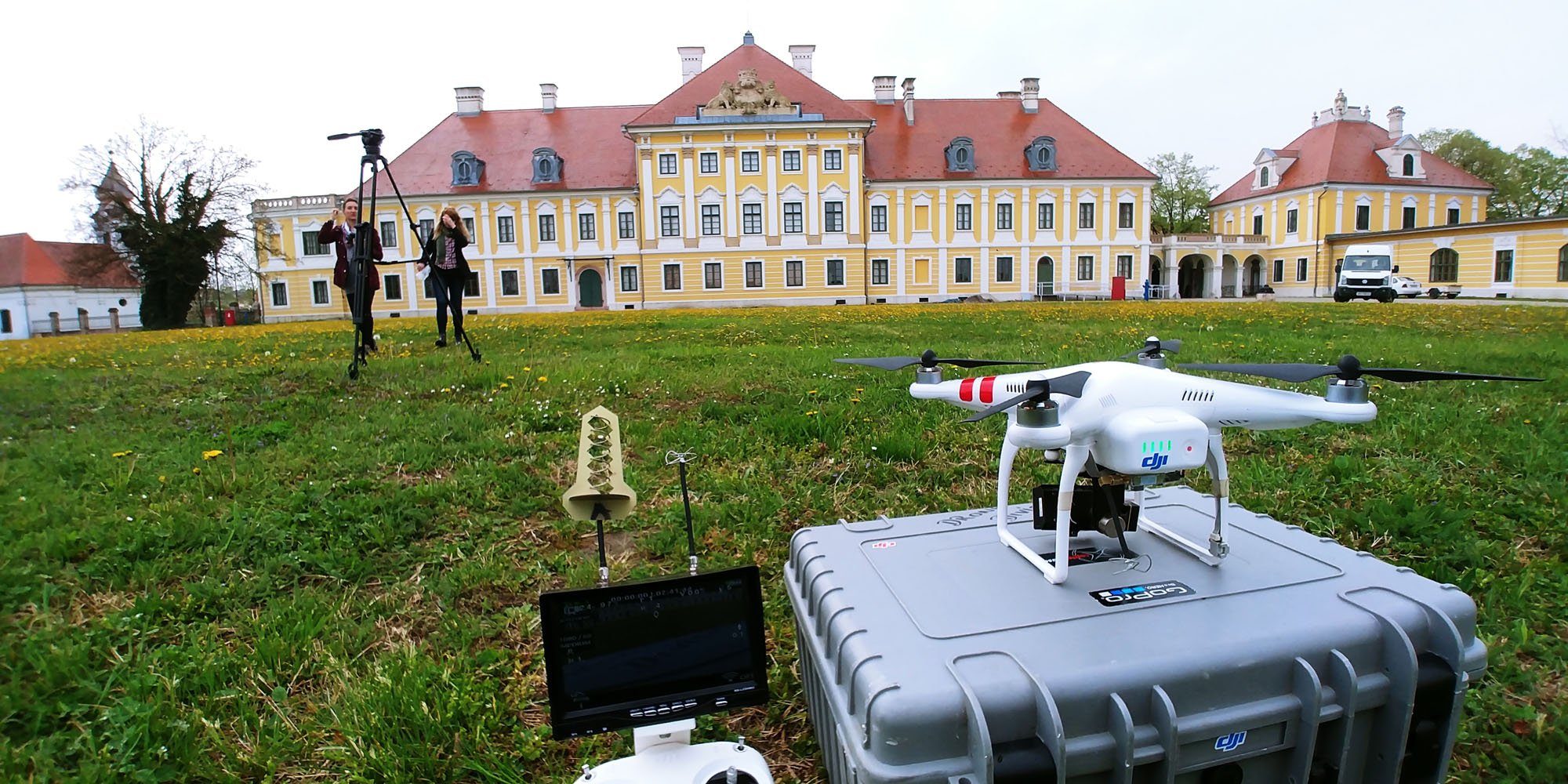 A large yellow and white historic building with red roof, two women with camera equipment filming in front, and a drone on a case in the foreground in a grassy area with small yellow flowers.