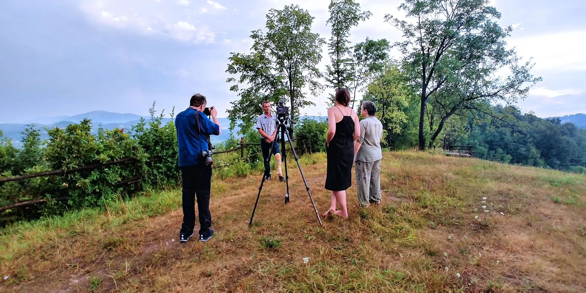 People recording a video on a grassy hillside with trees and mountains in the background.