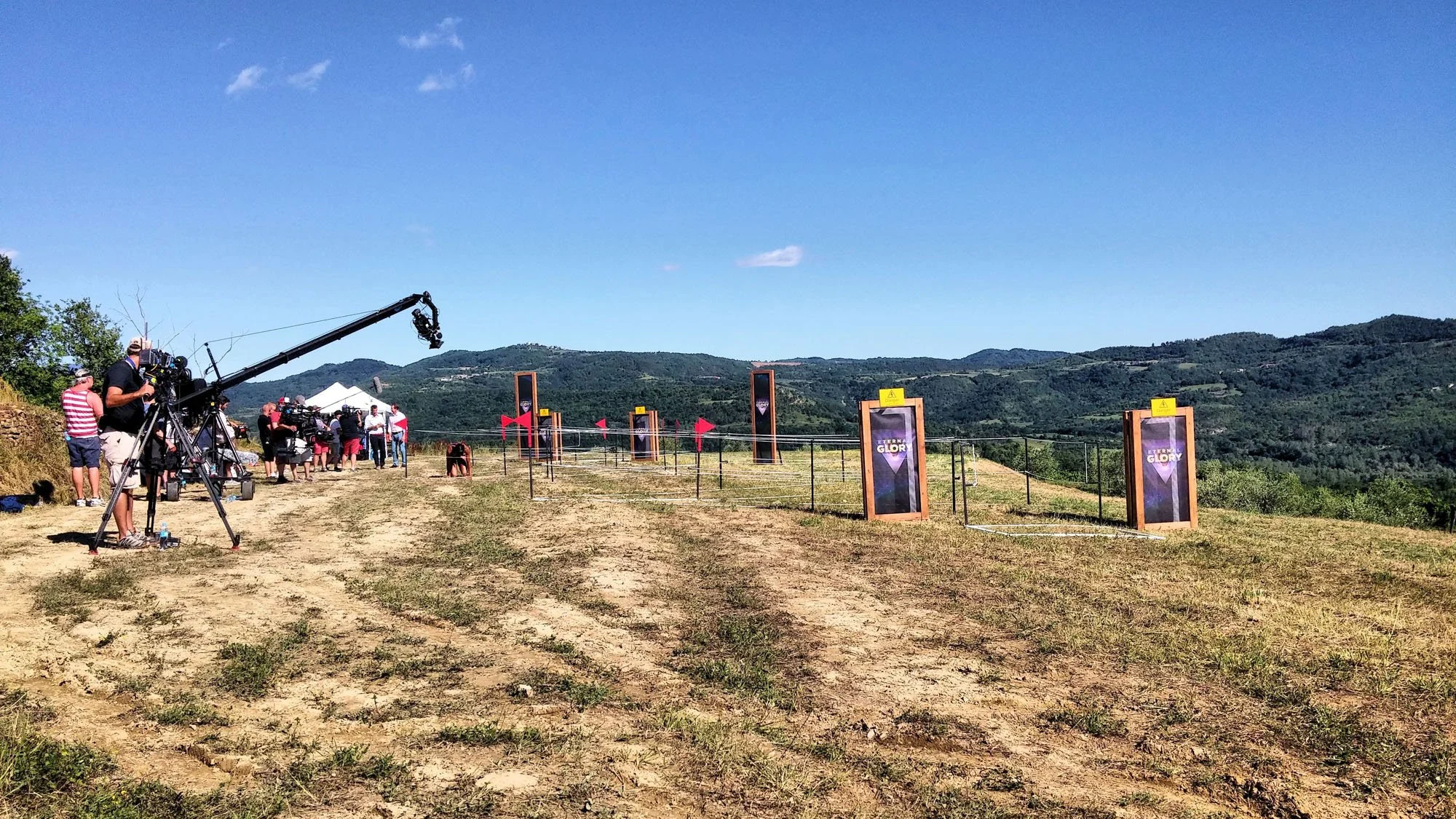 Filming crew setting up equipment for outdoor filming with mountains in the background, large vertical banners reading 'Eternal Glory', and a clear blue sky.