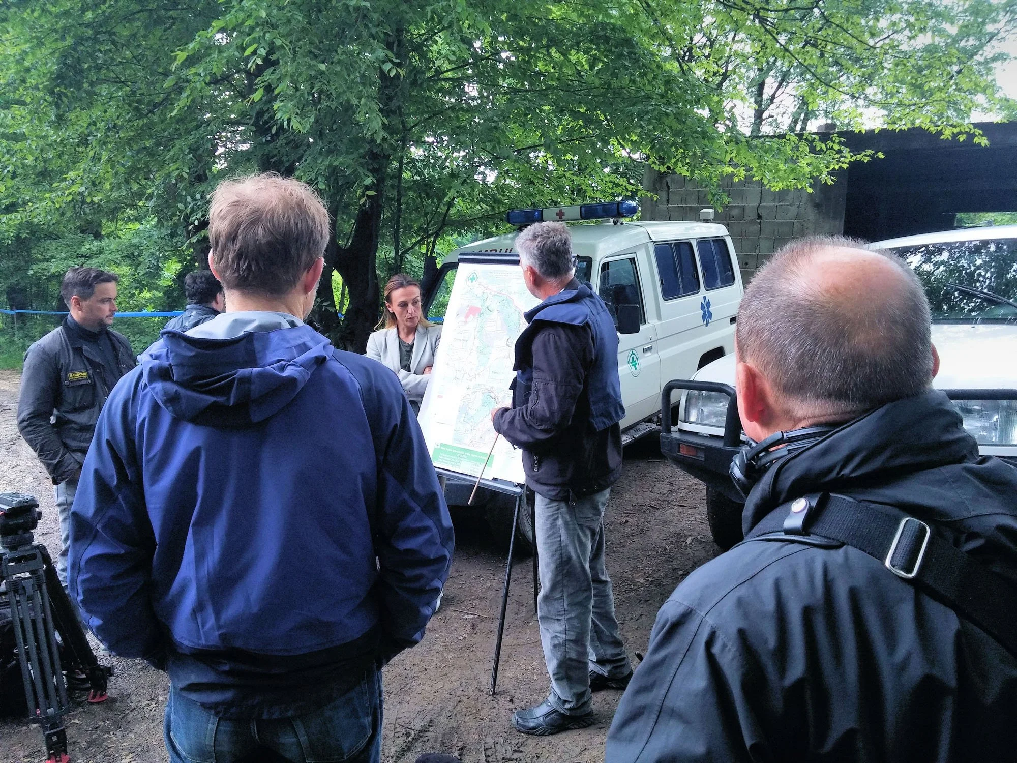 Group of people, including emergency responders, gathered outdoors under trees, discussing around a displayed map near police and emergency vehicles.