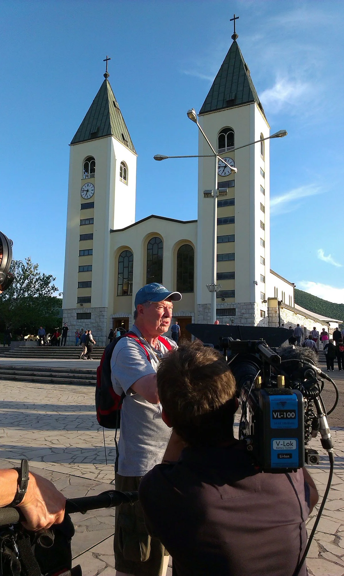 A filming crew is filming in front of a large church with two tall towers topped with crosses. The church is white with arched windows. Several people are walking around the plaza, and a man is standing near a camera operator.