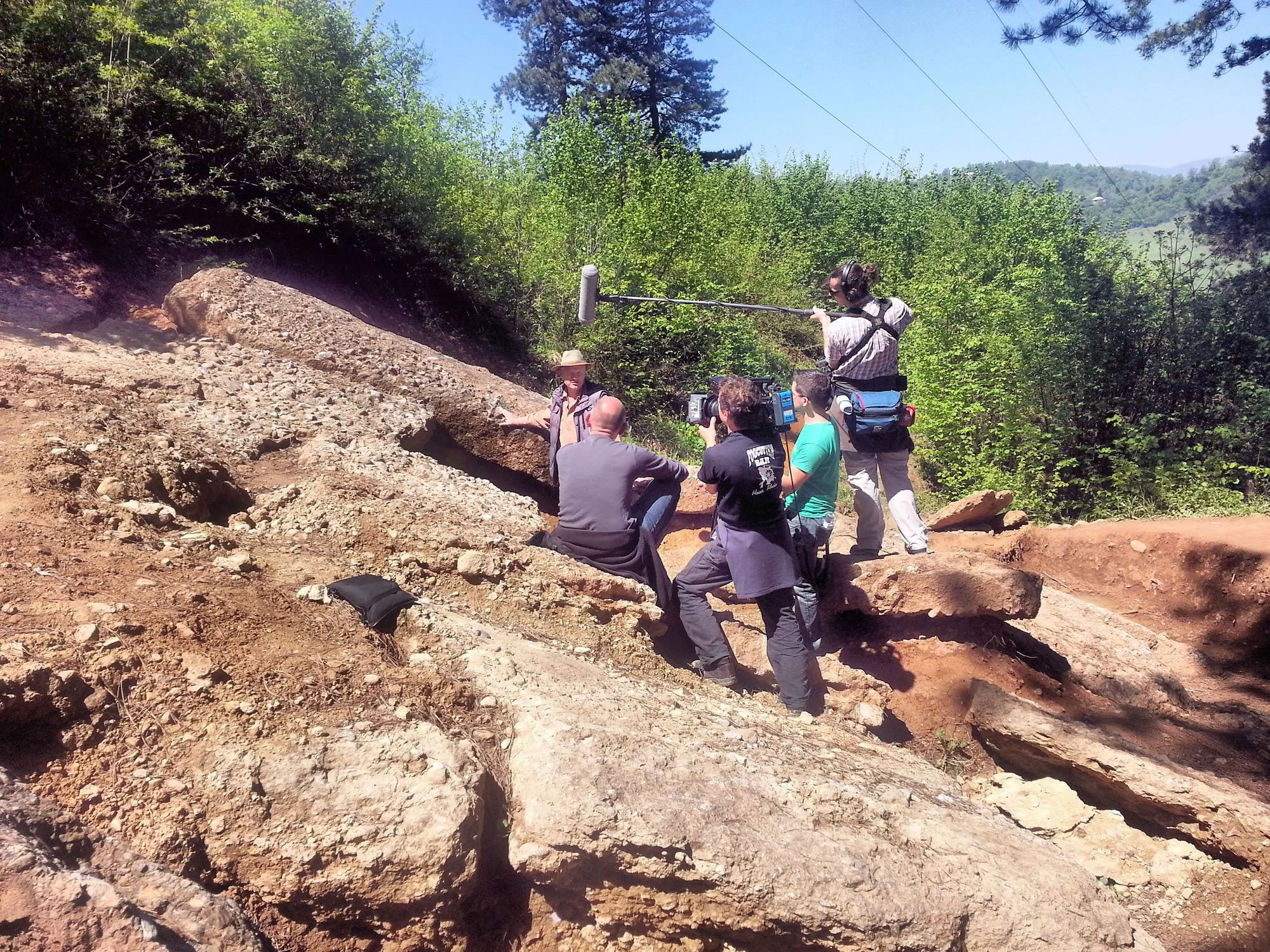 A group of people filming an outdoor scene on a mountainside with rocks and dense green trees under a bright blue sky.