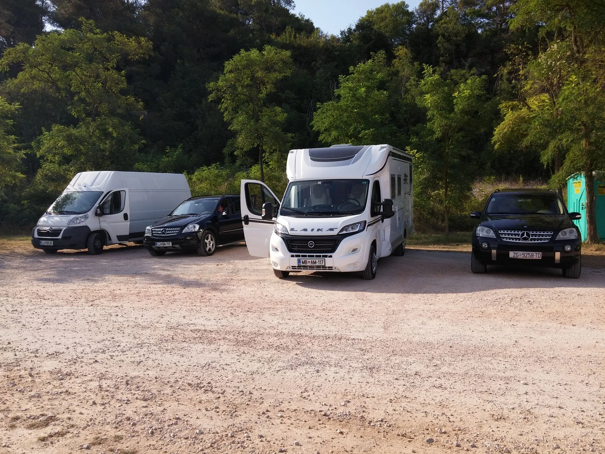 A row of five parked vehicles including a white camper van, two black cars, a white van, and a black SUV, in a dirt lot in front of a wooded area with green trees and a portable toilet on the right.