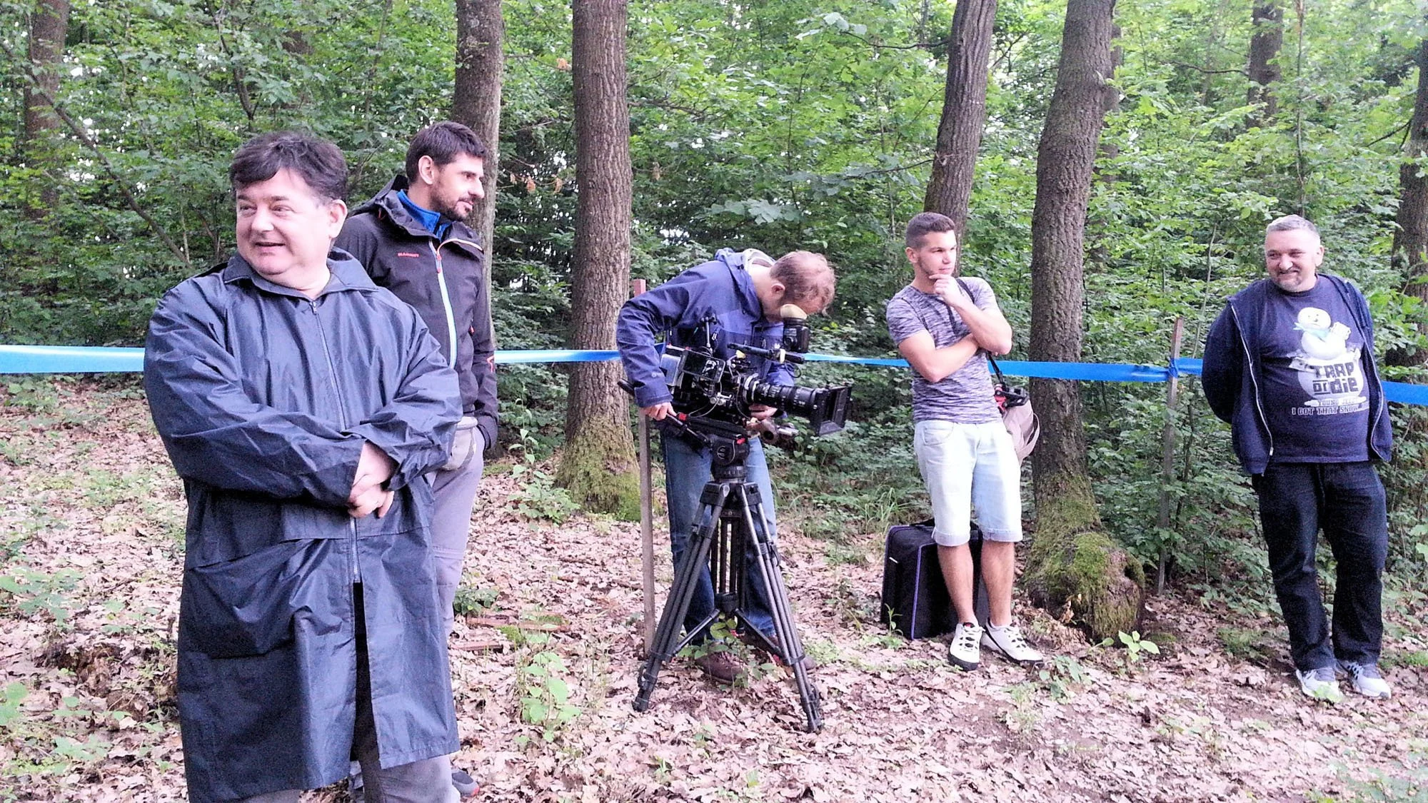 Group of five men standing in a wooded area with a camera on a tripod, some with backpacks, looking in different directions, in a forest setting.