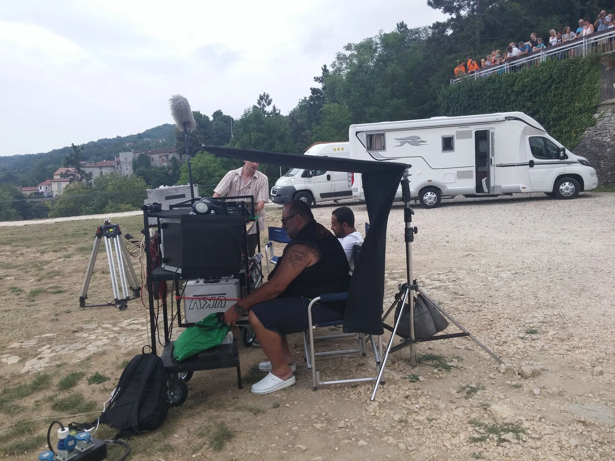 Filmmaking crew setting up equipment outdoors with a large crowd watching from a balcony above
