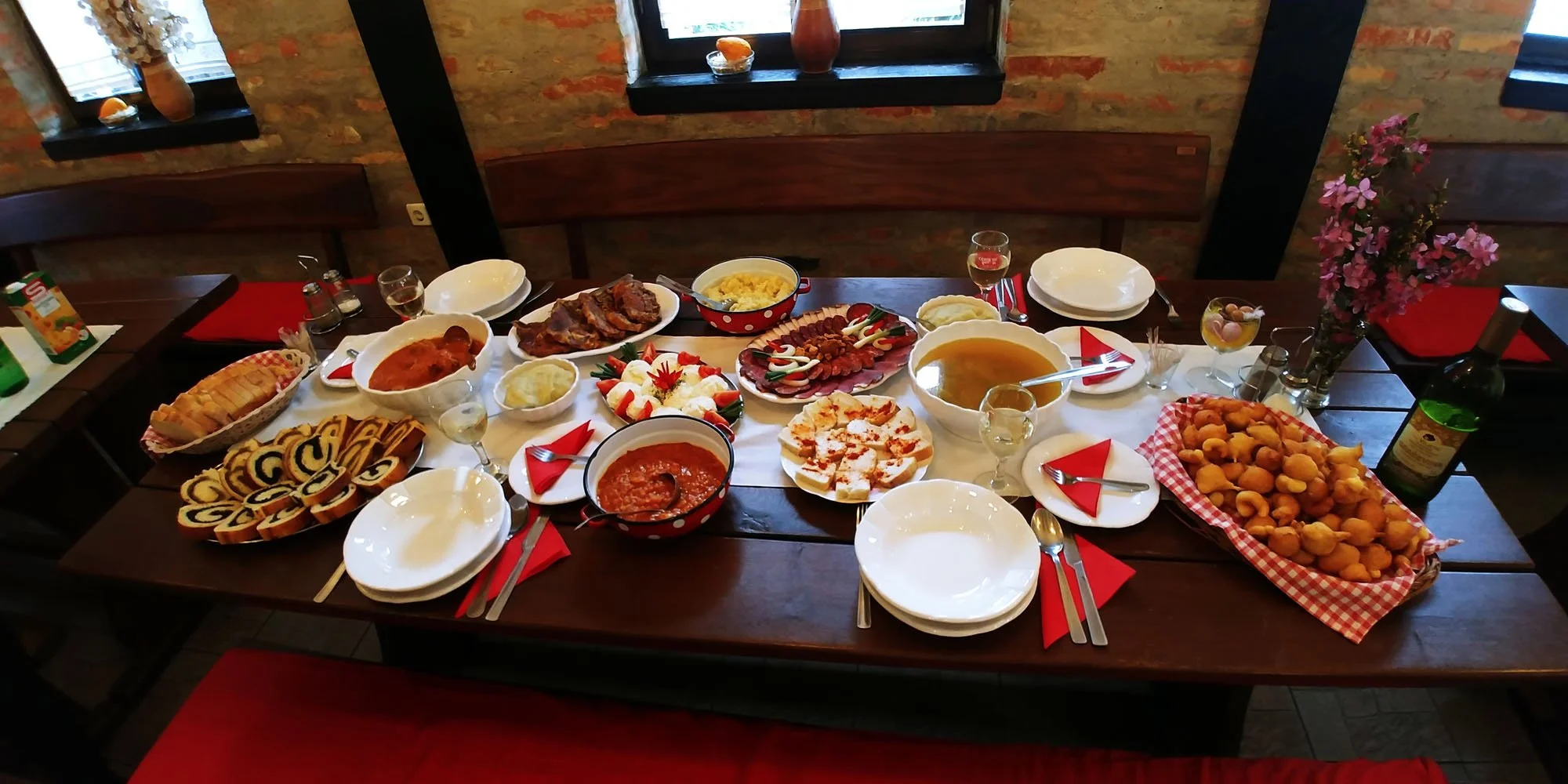 A large dining table set for a meal, filled with various dishes including salads, meats, bread, and desserts, with wine glasses and silverware, in a rustic restaurant with brick walls and small windows.