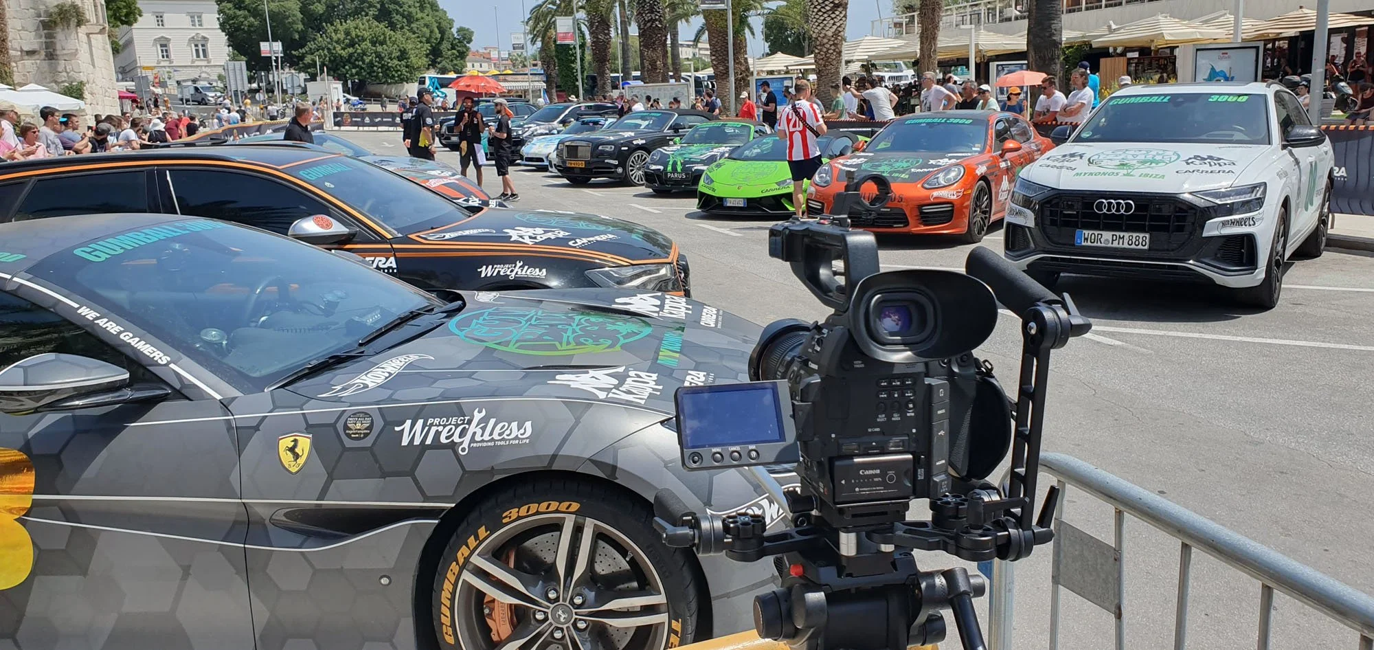 A car show with various sports cars and luxury vehicles parked outdoors, people walking and observing, and a professional camera setup in the foreground.