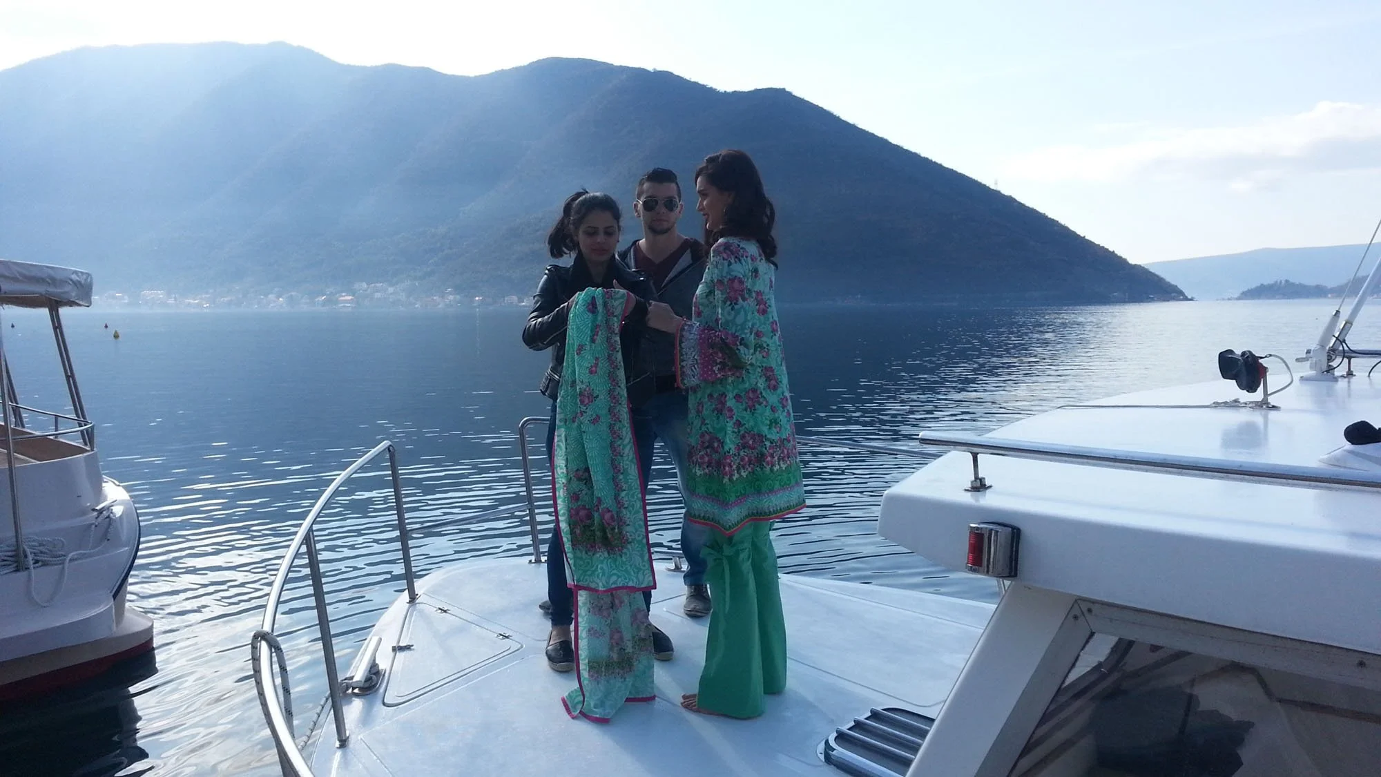 Three people stand on a boat deck overlooking a body of water with mountains in the background. One woman is dressed in traditional colorful clothing, while the other woman wears a dark leather jacket. A man in sunglasses stands between them, looking