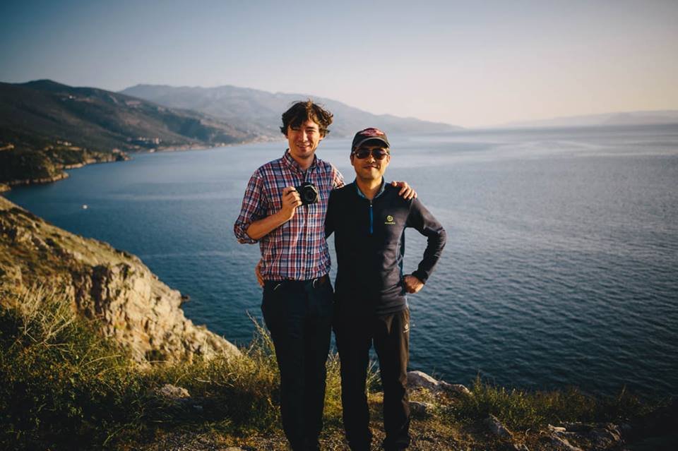 Two men standing on a hill overlooking a large body of water with mountains in the background. One is holding a camera.