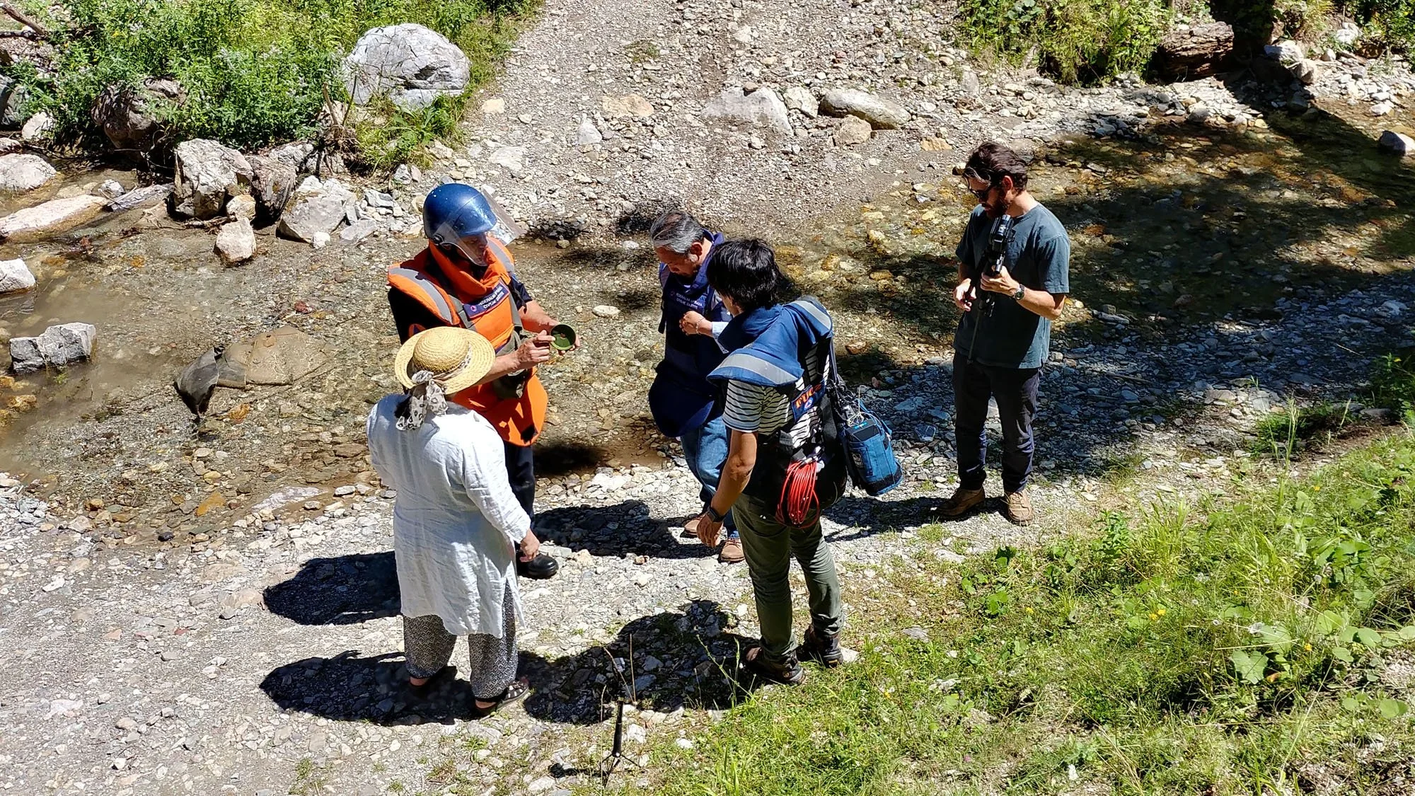 Group of people outdoors by a creek, engaging in a conversation or activity, with some wearing outdoor gear and equipment.