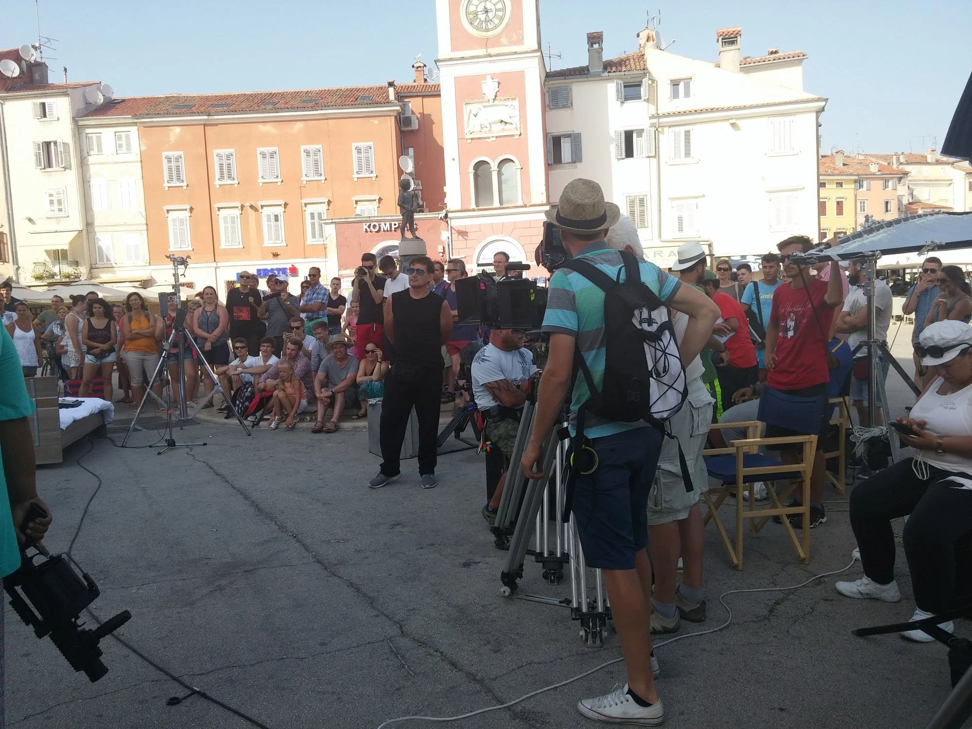 A crowd of people gathered outdoors in front of colorful buildings with shutters, during what appears to be a sunny day. There are cameras and filming equipment set up, suggesting a filming event or street performance. A man in a hat is operating a c