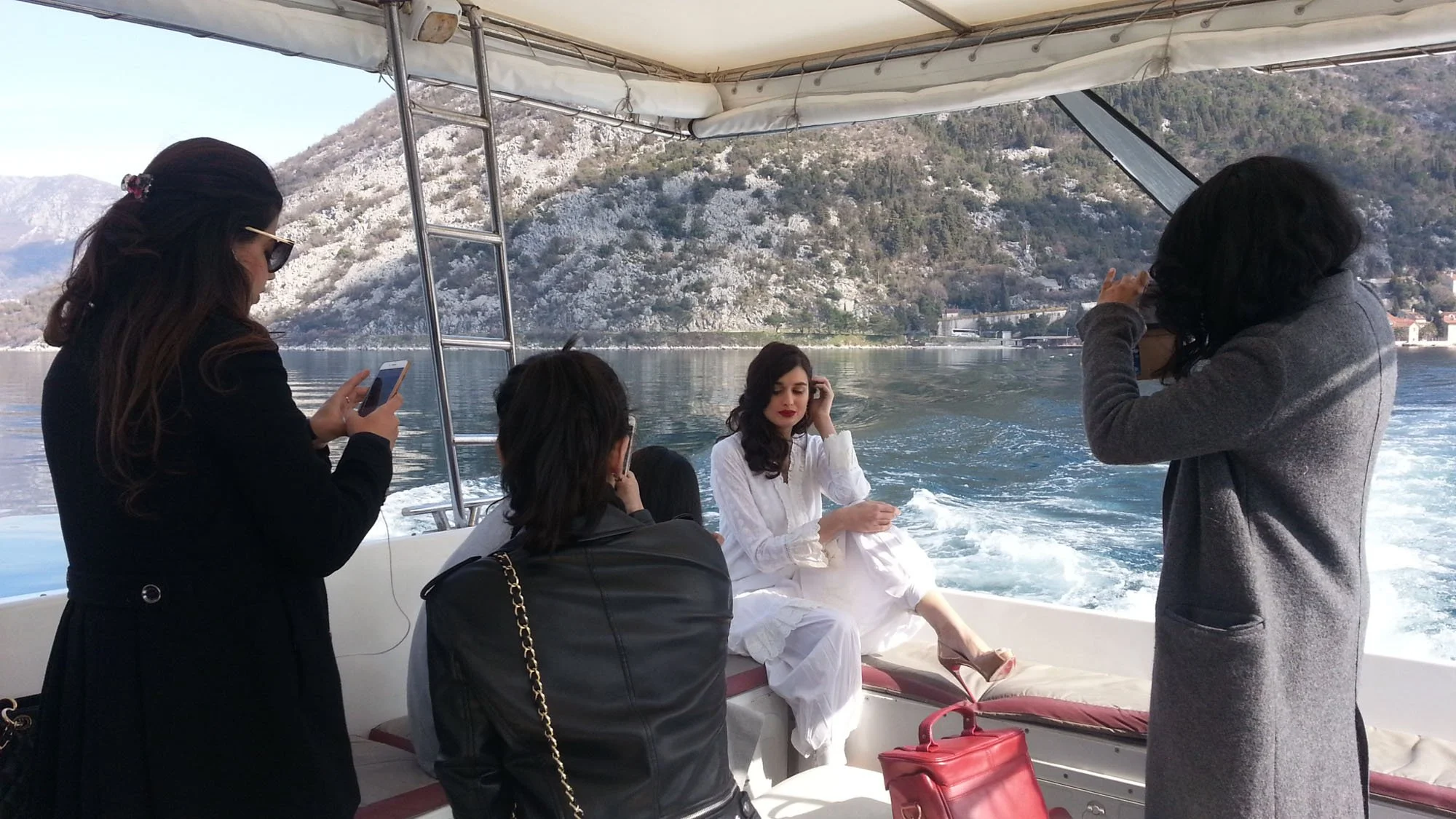 A woman in a white dress sitting on a boat with a scenic mountain background, surrounded by four other women taking photos and talking.