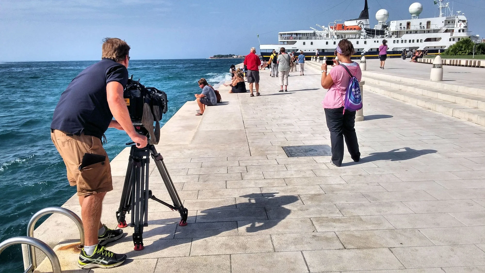 People on a pier near a cruise ship, some taking photos, with the ocean on the left and the ship docked on the right.