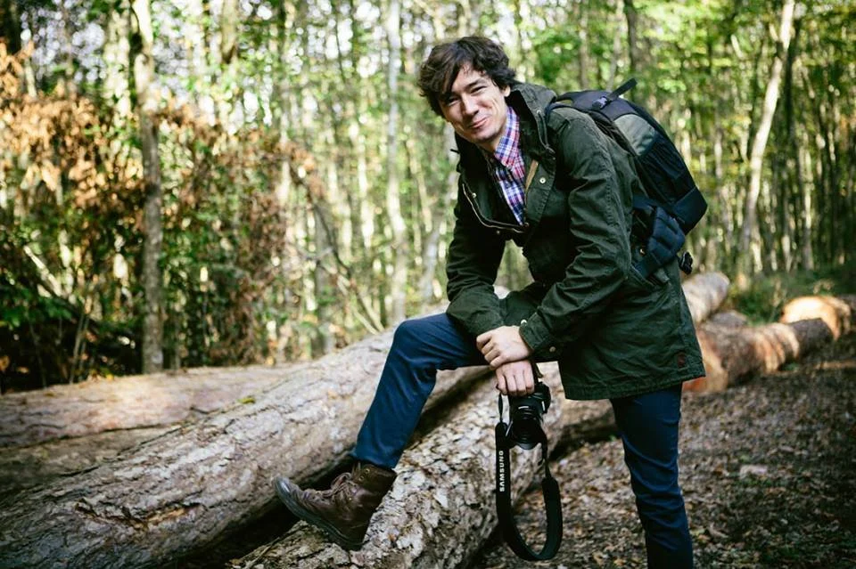 A man with dark hair and a plaid shirt, wearing a green jacket and jeans, is crouching on a fallen log in a forest. He has a backpack and is holding a camera.
