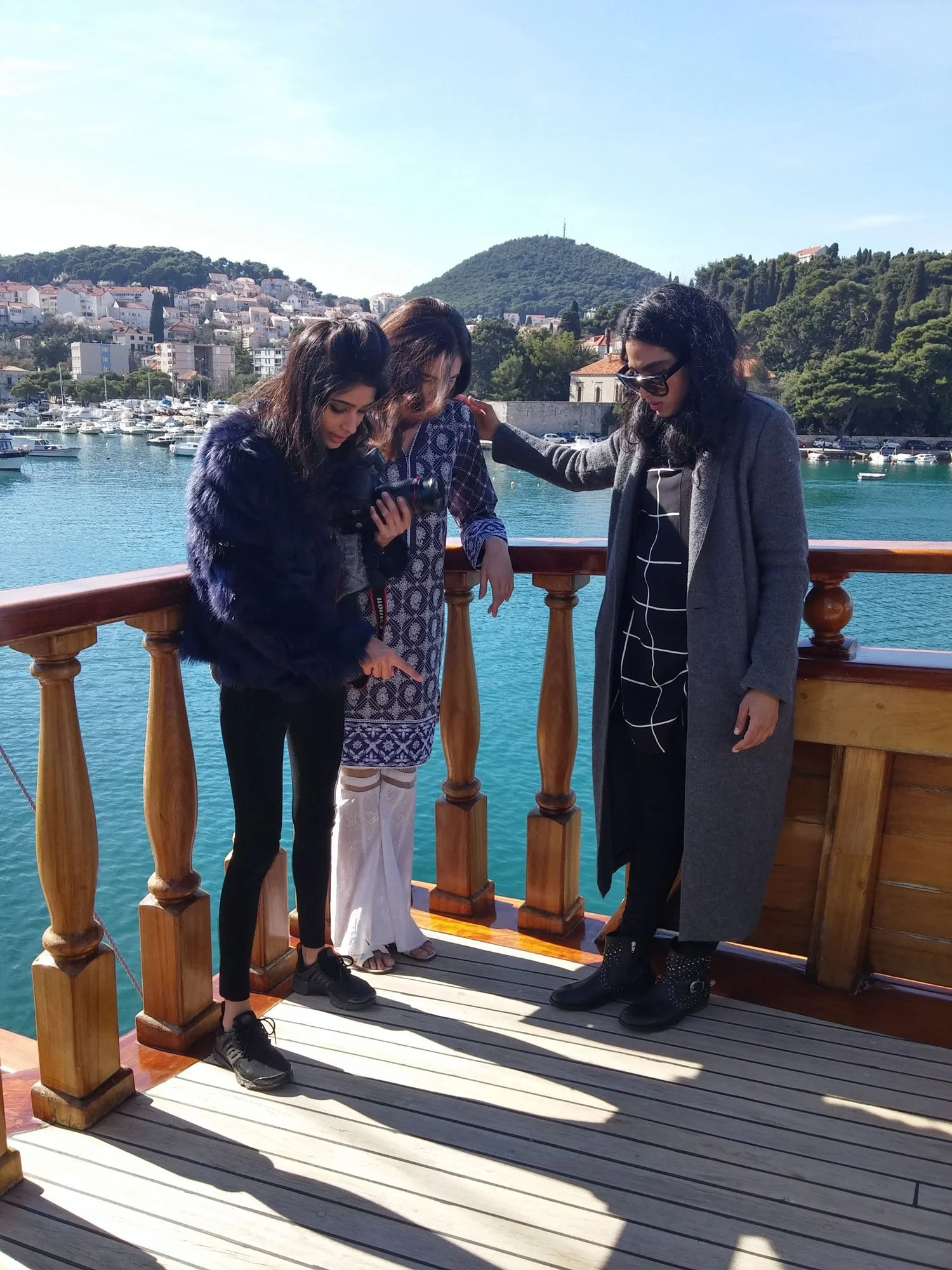 Three women standing on a boat's deck with a marina and hillside in the background, looking at a camera and pointing at something.