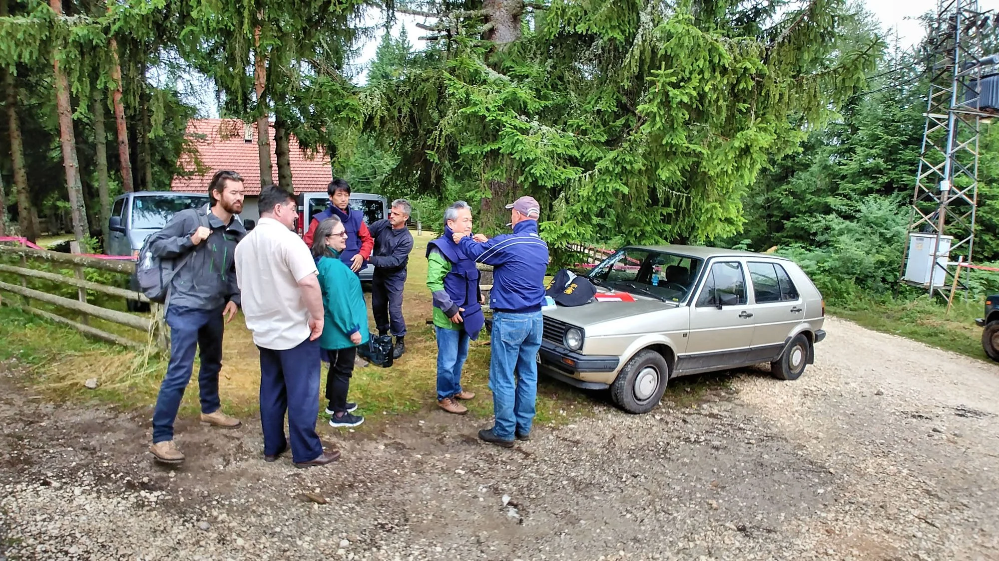 A group of people gathered outdoors in a forested area around a small silver car. They appear to be having a discussion, with one person showing something to the group. Several cars are parked nearby, and there is a large green tree behind them.