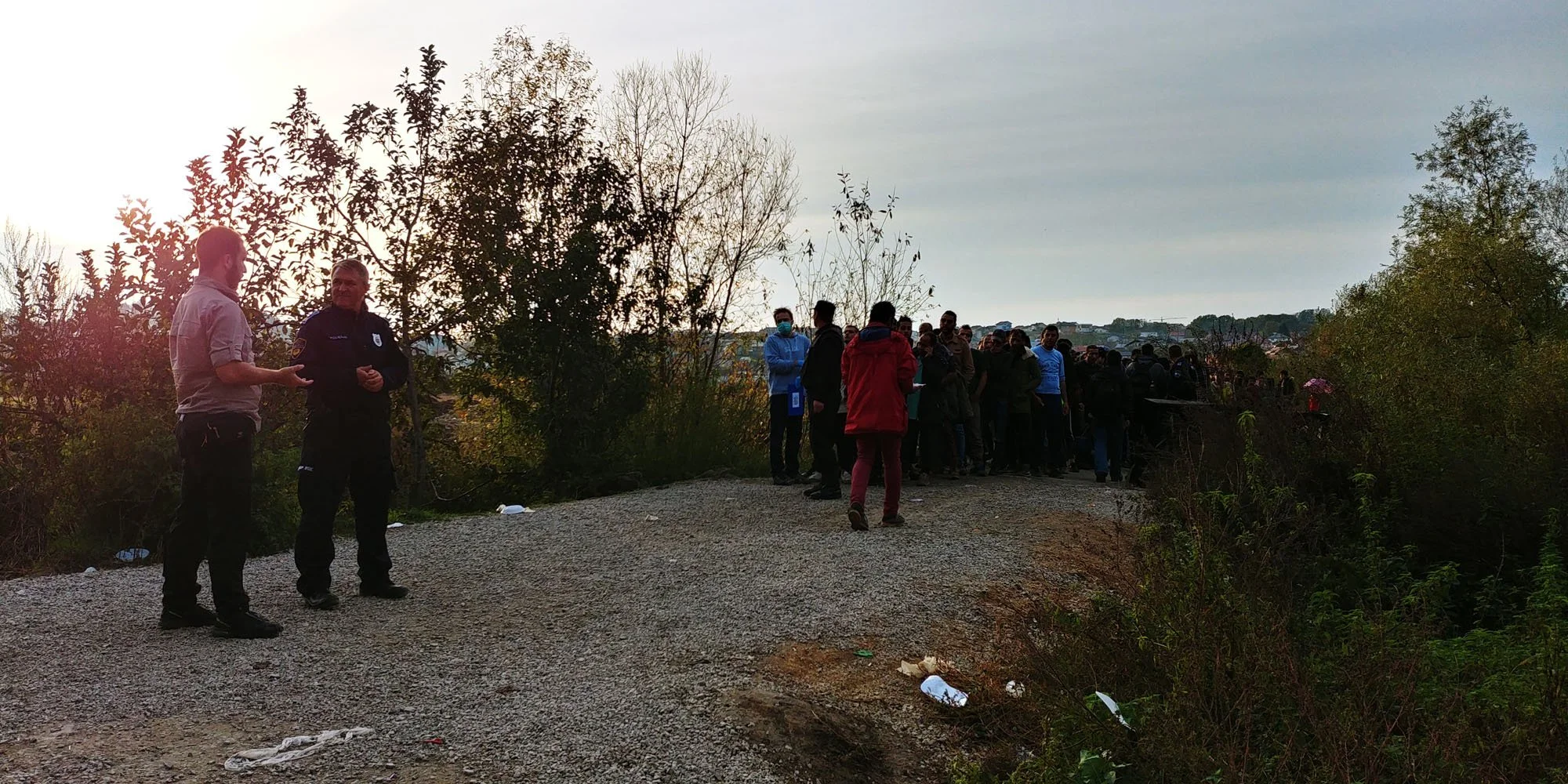 A group of people, including police officers, standing on a dirt path outdoors, with some people wearing masks and trees in the background.