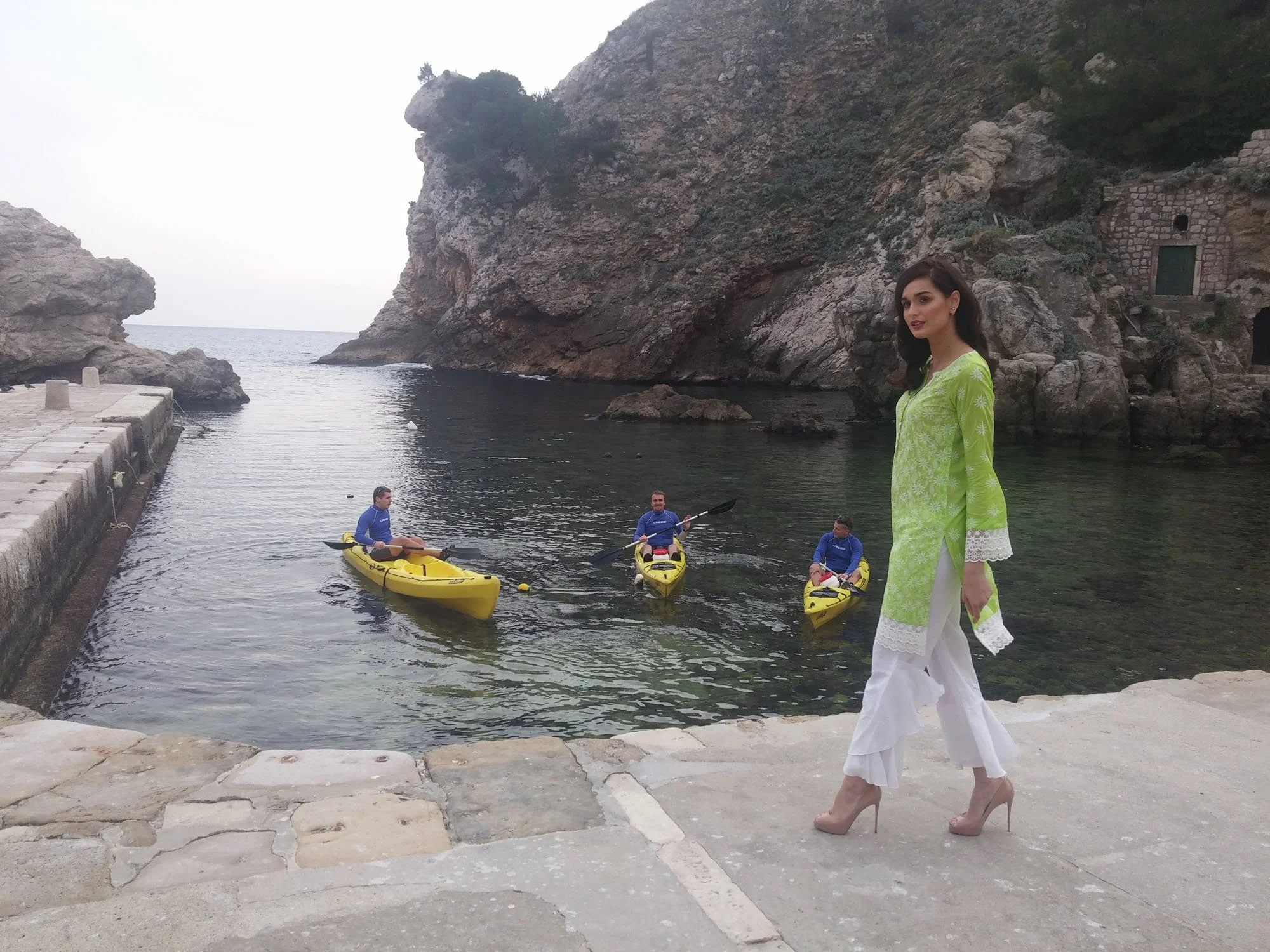 A woman in a bright green dress with white floral and lace details stands barefoot on stone paving near the water, with a scenic rocky coastline and three people kayaking in yellow boats in the background.