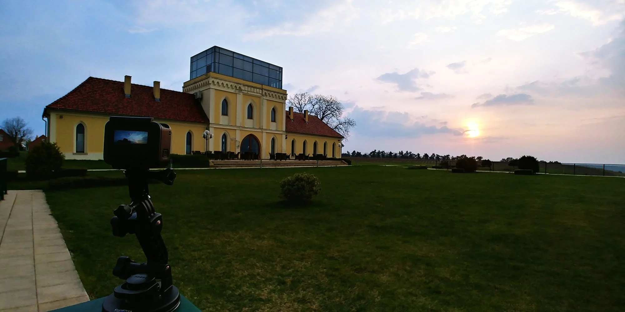 A yellow building with a glass rooftop structure in a lush grassy field during sunset, with a camera mounted on a tripod in the foreground.