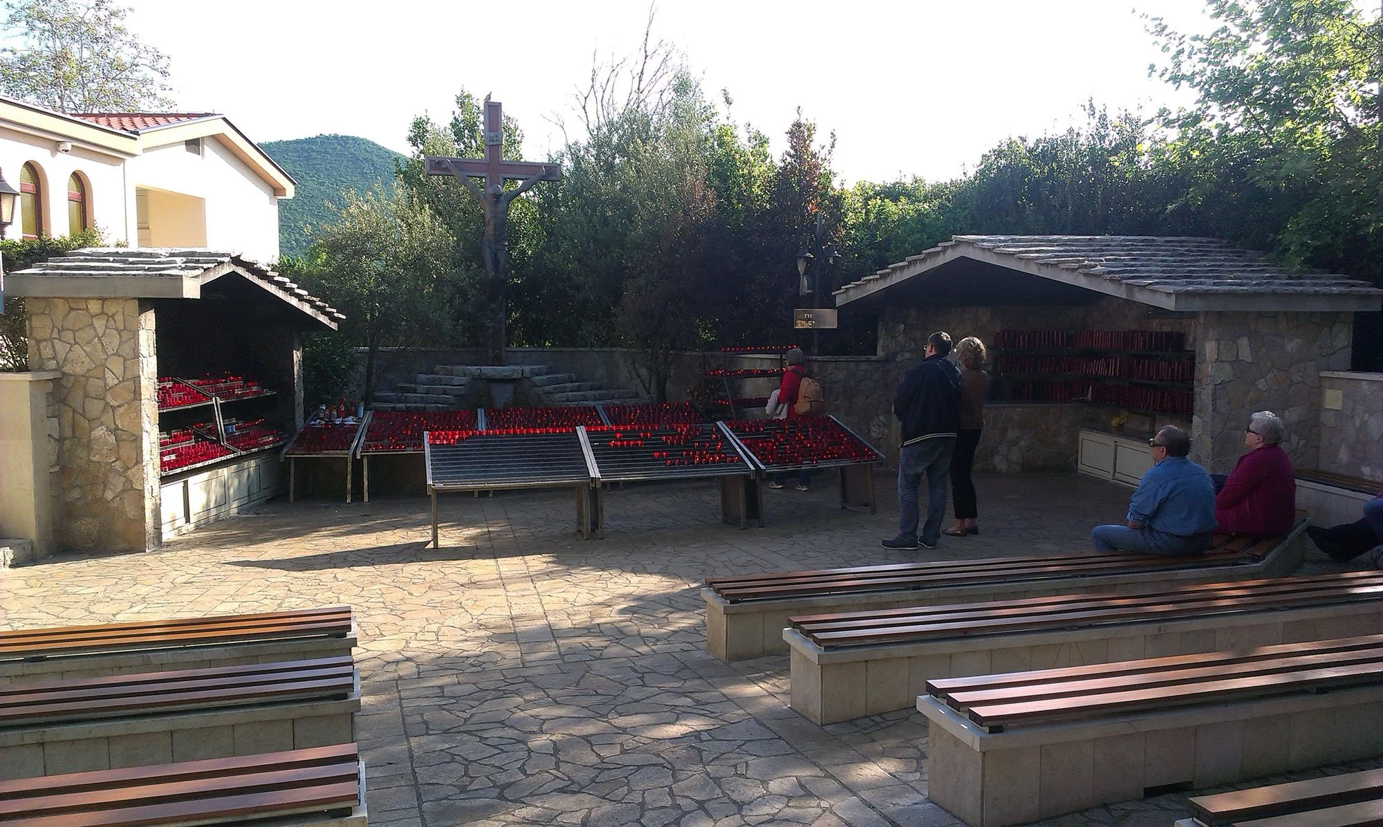 A small outdoor chapel with a crucifix with Jesus, surrounded by trees and mountains. People are sitting on benches or standing near the stone structures with red votive candles.