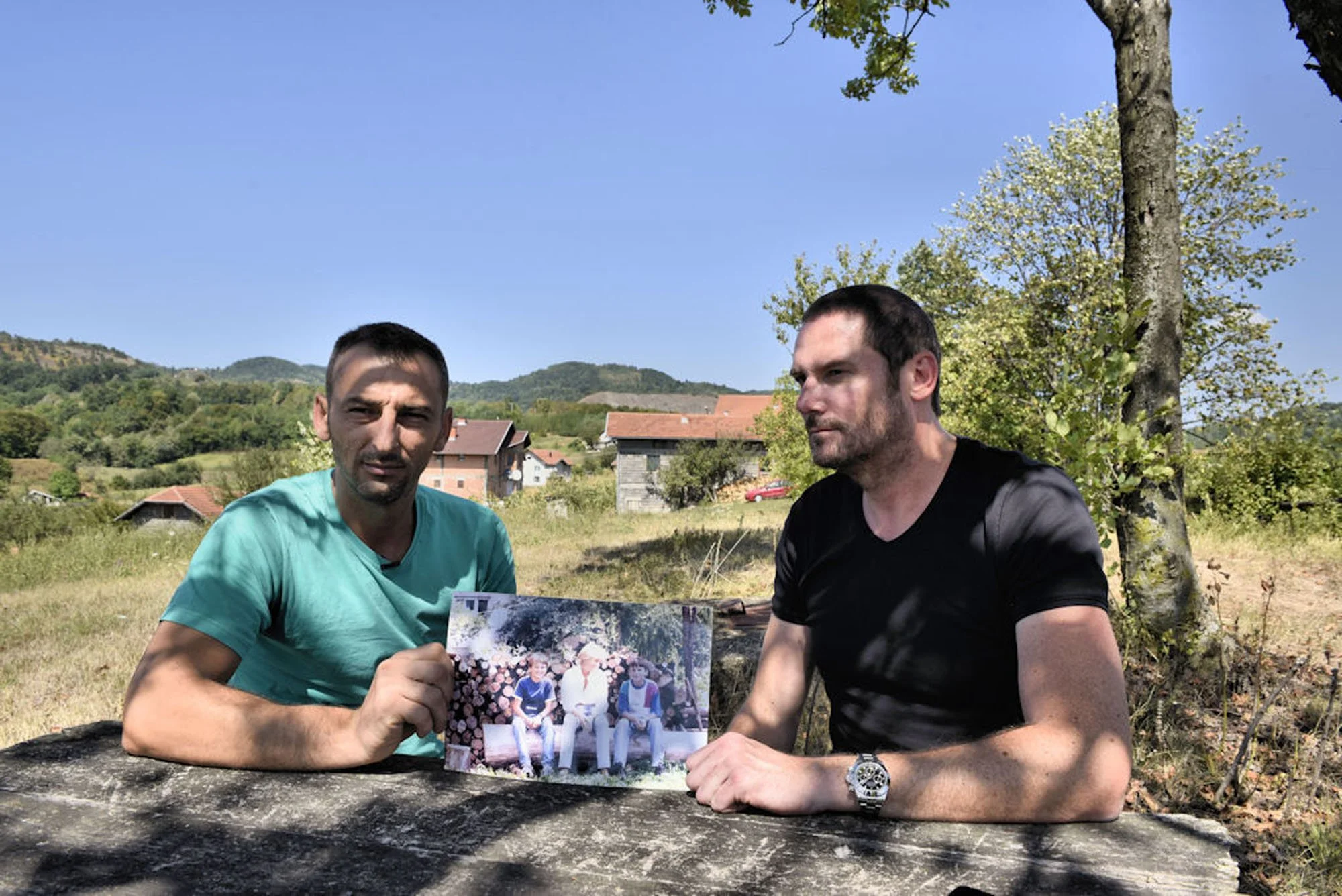 Two men are sitting outdoors at a wooden table, holding a photo album and a large photograph. The background features green hills, trees, and a few houses under a clear blue sky.
