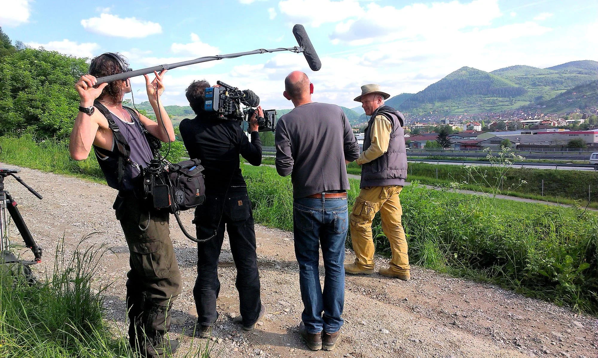 A film crew filming an interview outdoors in a scenic mountainous area with multiple crew members, including a person holding a boom microphone, a camera operator, and two men in conversation, one wearing a broad hat and vest.