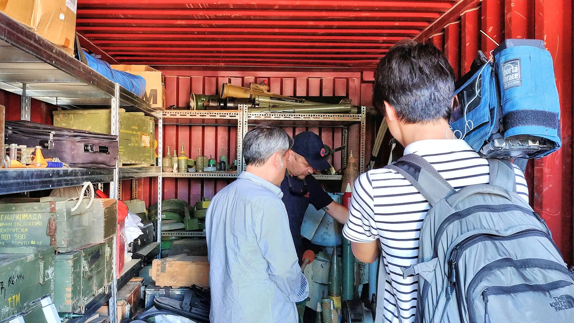 Three men in a workshop or storage area with shelves holding various tools and military equipment, including rocket launchers and ammunition, as a fourth person looks at a map or document.