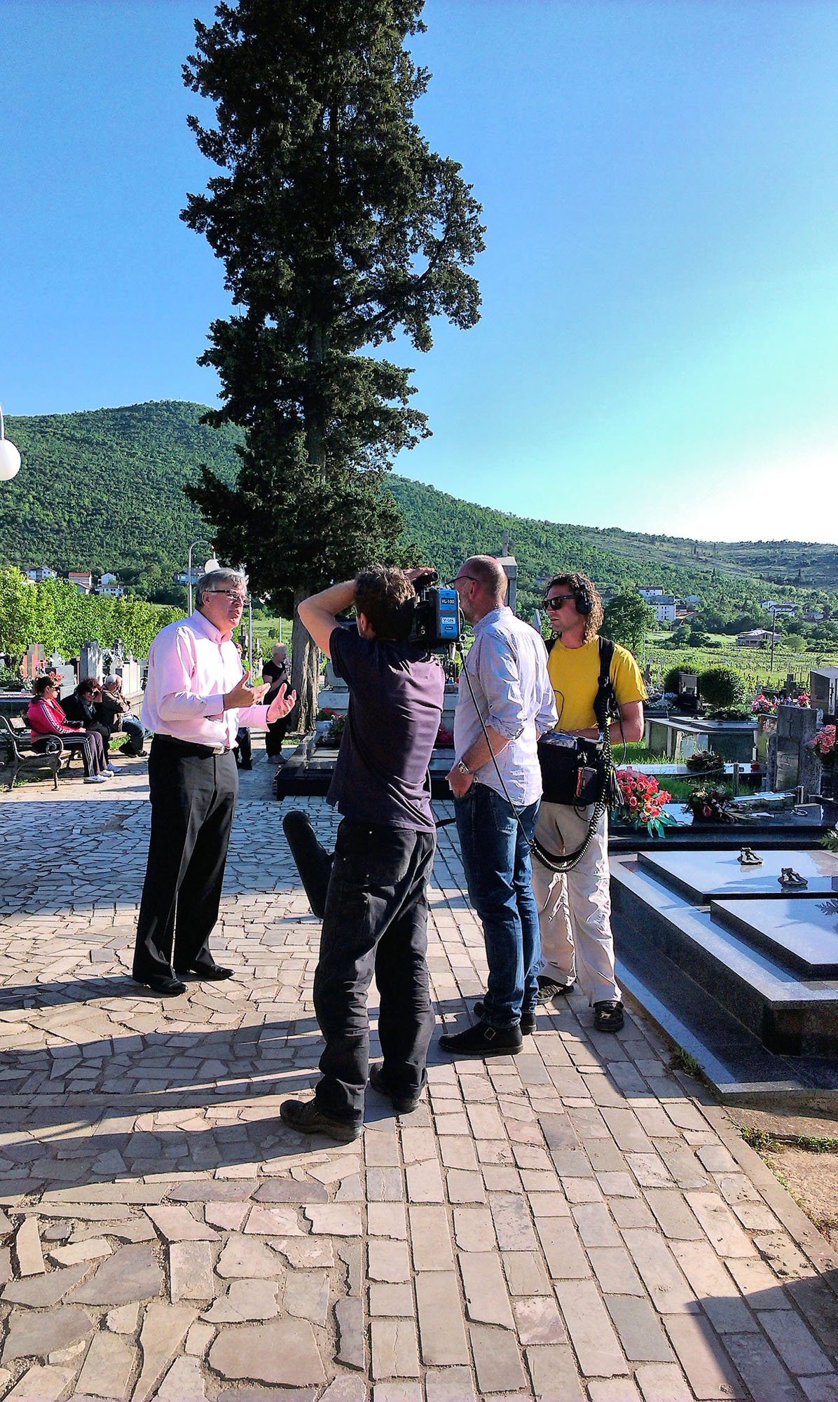 People filming a man speaking in a cemetery, with a large tree, green hills, and graves visible in the background.