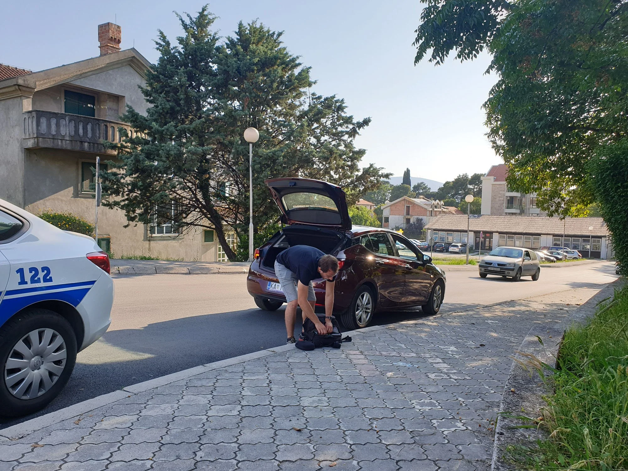 A man is kneeling next to a parked black car with its trunk open, on a parking lot with cars and buildings in the background, and a woman is looking into a backpack on the ground in front of him.