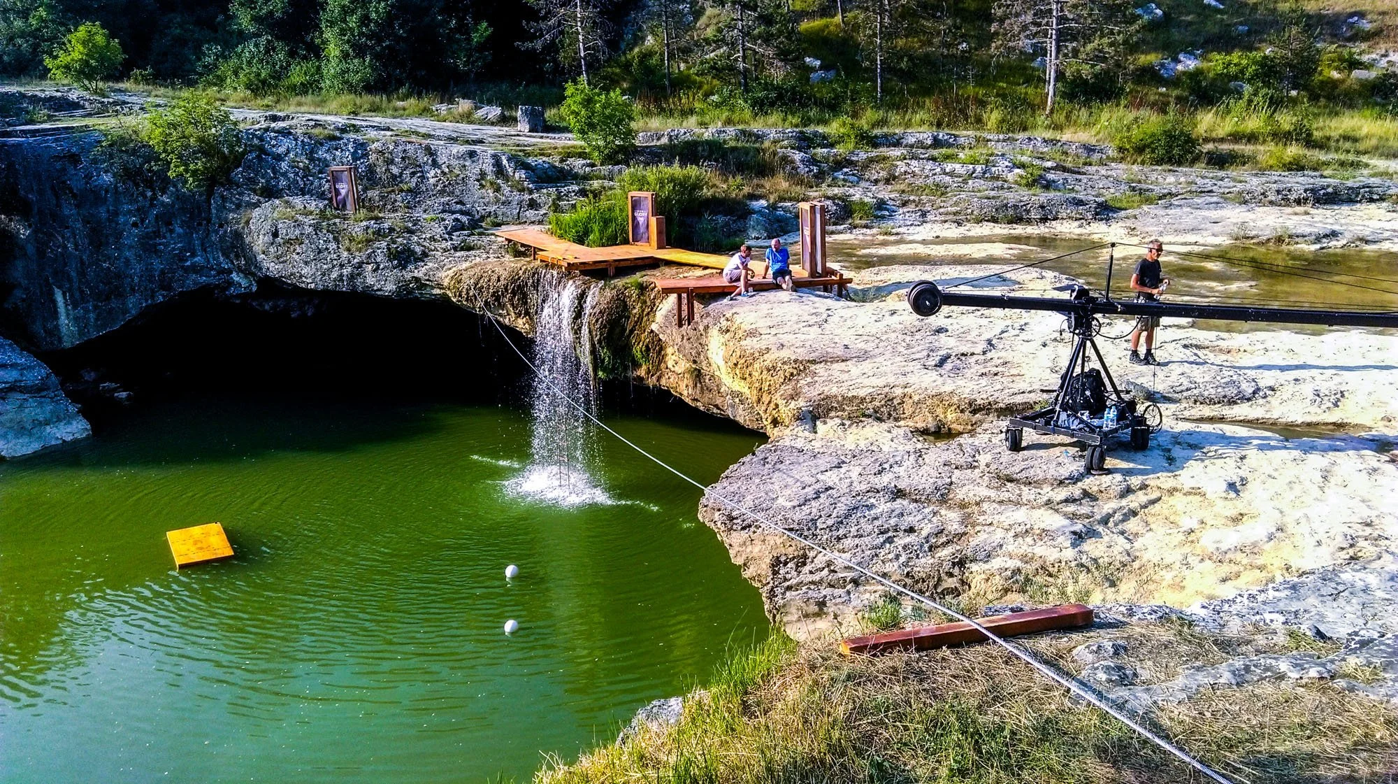 People fishing at a rocky outdoor location with a small wooden platform and a camera on a wheeled platform, overlooking a green water pool with a waterfall