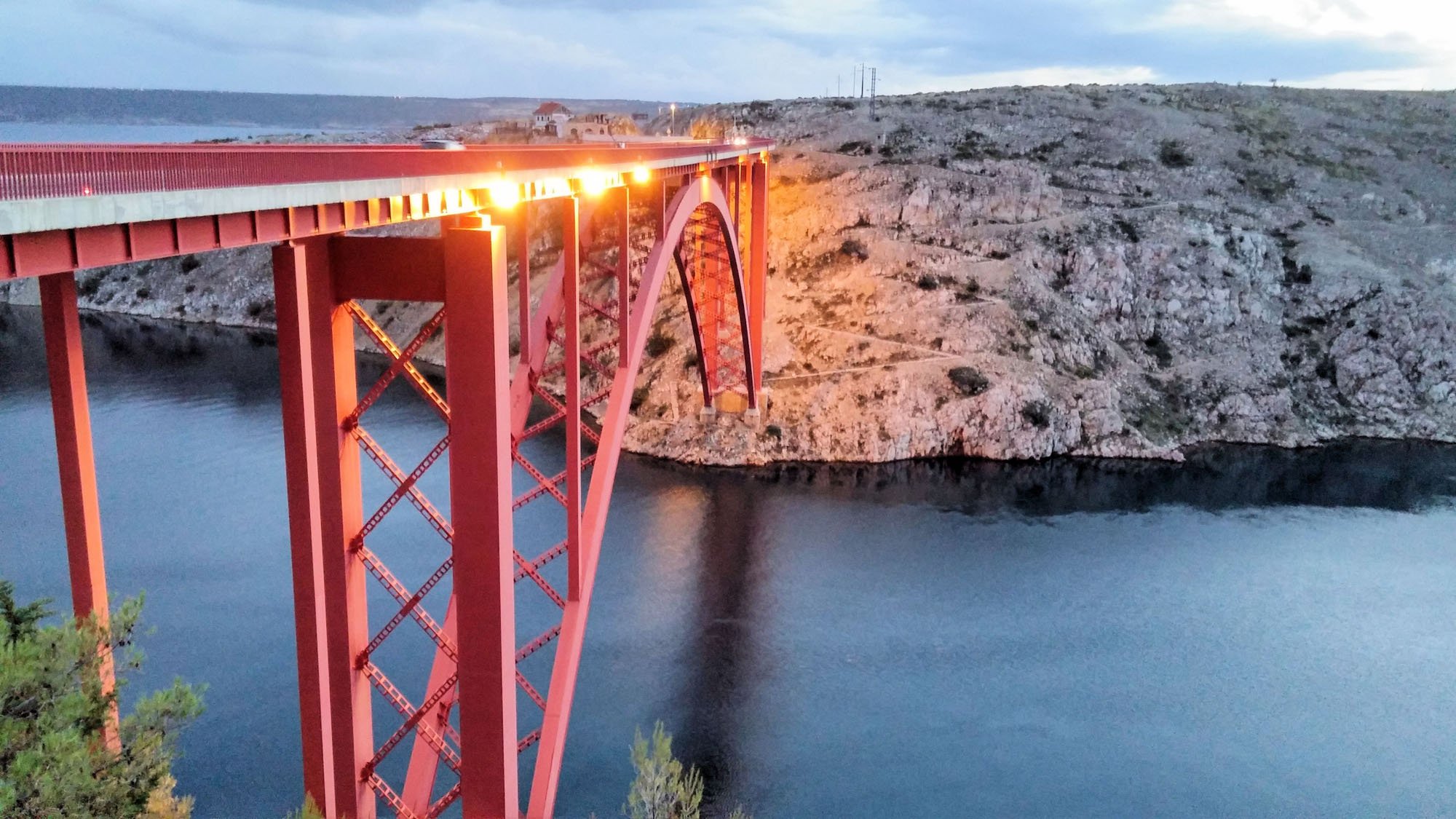 A large red bridge spanning over a body of water with rocky hills and a cloudy sky in the background, illuminated by lights.