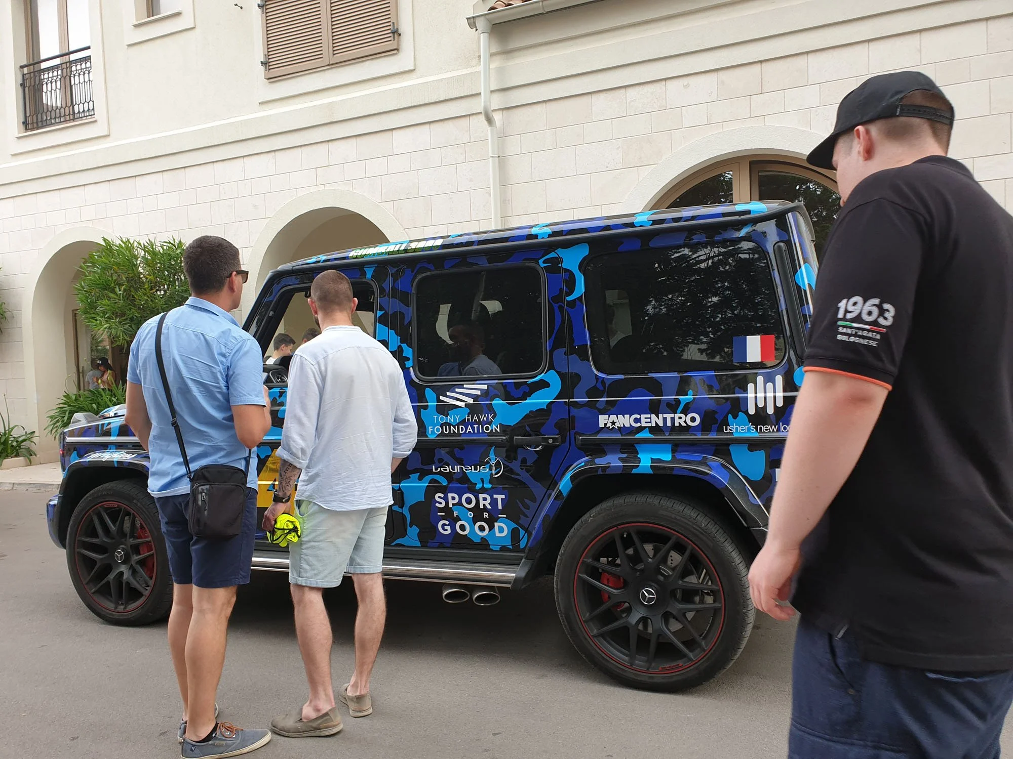 People gathered around a blue camouflage-patterned vehicle with logos related to sports and a foundation, parked on a city street in front of a beige building with arched windows. One person wears a black cap and black shirt, while others are dressed
