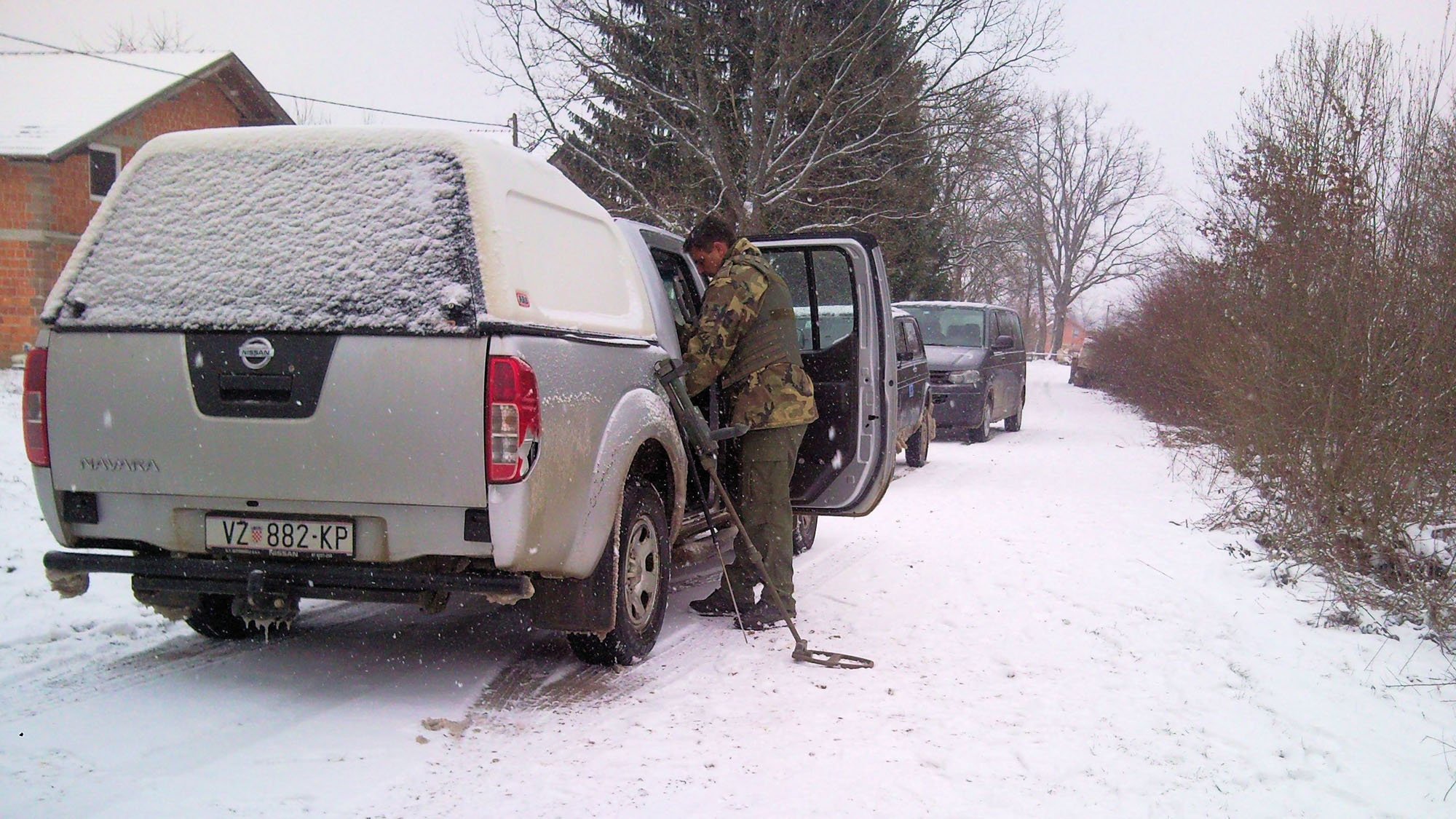 A person using a metal detector near a white Nissan pickup truck covered in snow outside on a snowy street, with trees and other vehicles in the background.