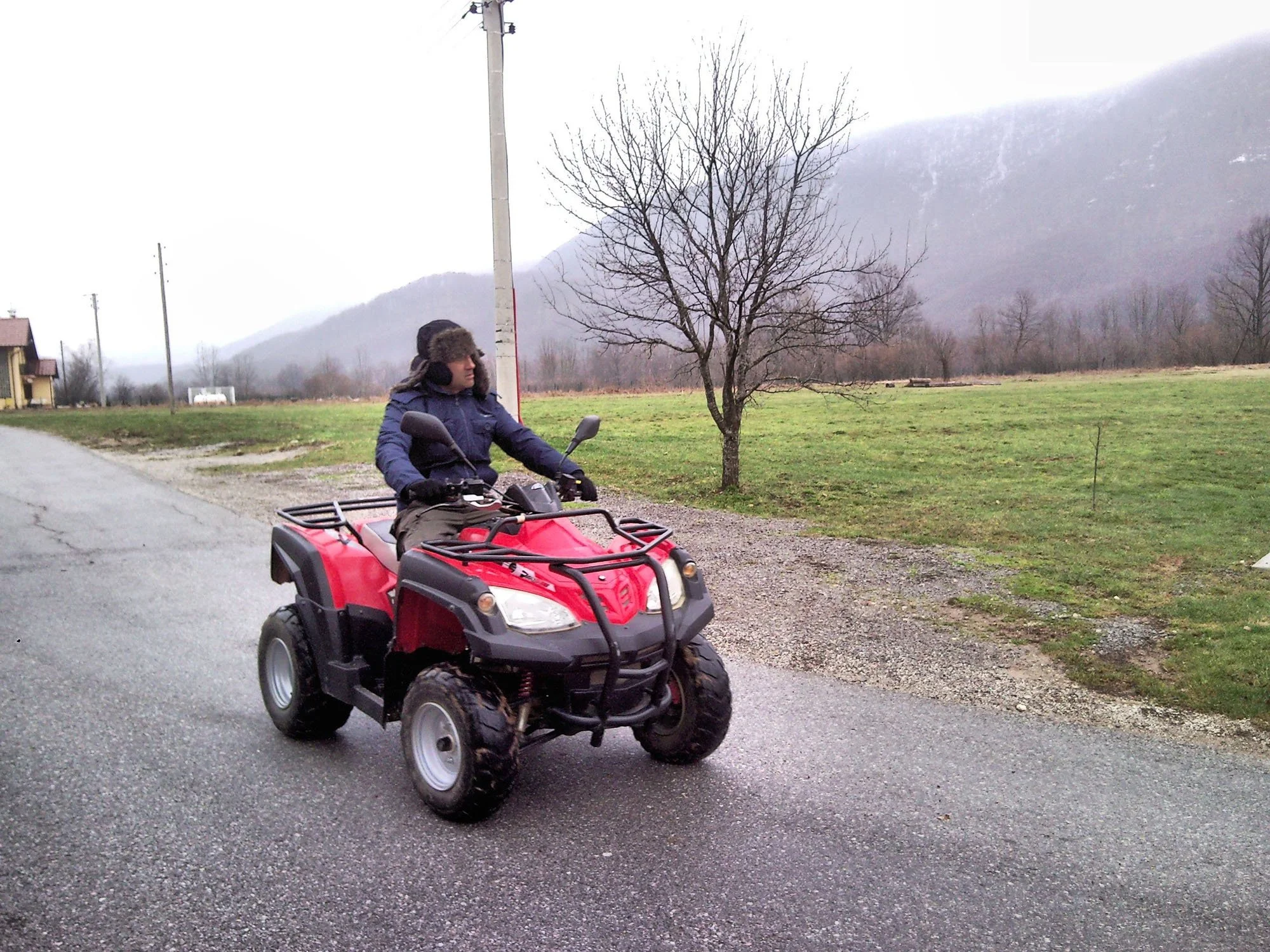 A person wearing winter clothing and a hat riding a red all-terrain vehicle (ATV) on a rural road with grassy fields and leafless trees, mountains in the background, and a cloudy sky.