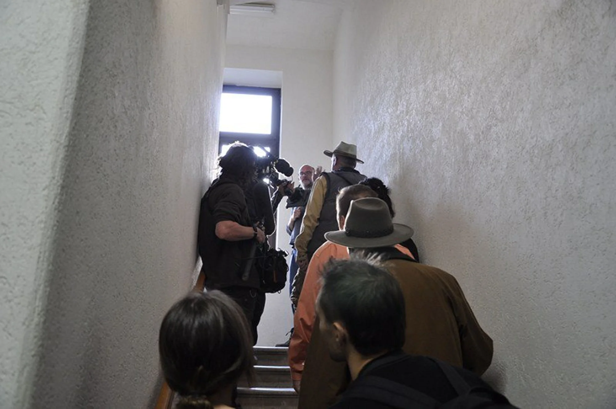People standing and waiting in a narrow, stairway hallway with beige textured walls, sunlight coming through a window at the top of the stairs, some wearing hats and backpacks, one person is filming or taking a picture.