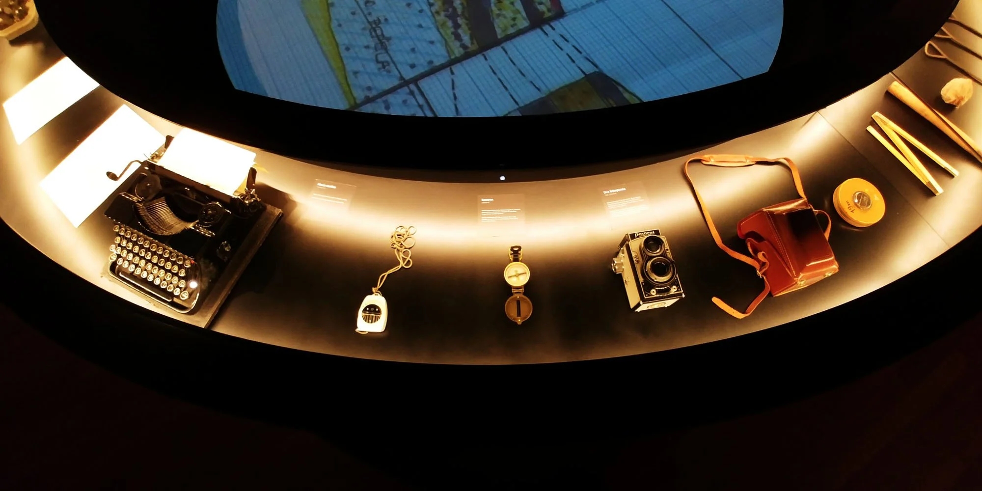 Display of vintage and electronic items, including a typewriter, camera, and bag, on a curved black surface in a museum exhibit.