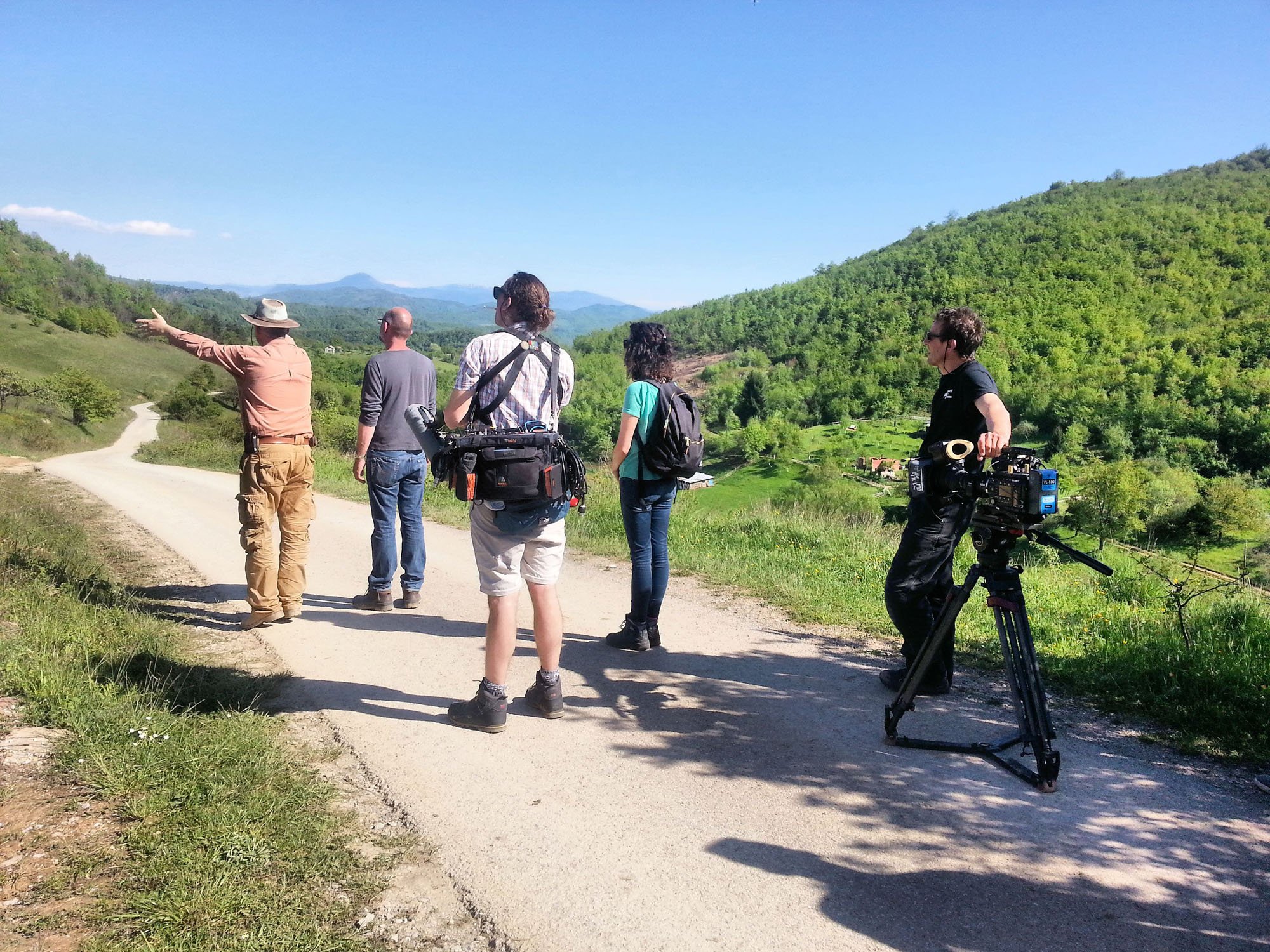 Group of people, including a man with a hat, a man with sunglasses, a woman with a backpack, and a man operating a large camera on a tripod, standing on a dirt road in a hilly, green landscape under a clear blue sky, with mountains in the distance.