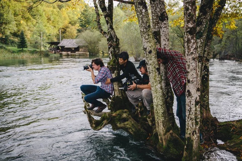 Four friends sitting by a river, with two of them taking photos and one peering over the water, surrounded by trees in a forested area.