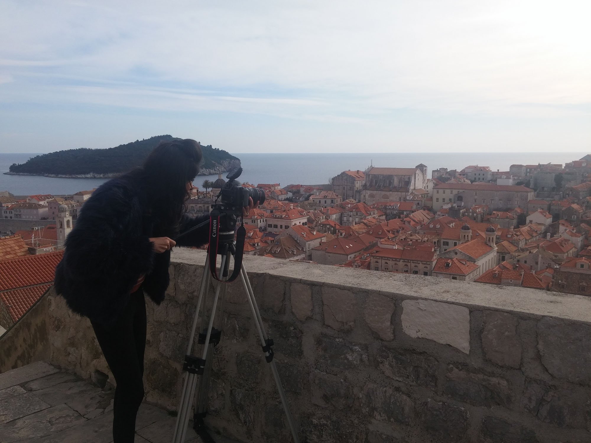 A woman with dark hair taking photos with a camera on a tripod overlooking a city with red-tile roofs, a church, and the ocean in the background.