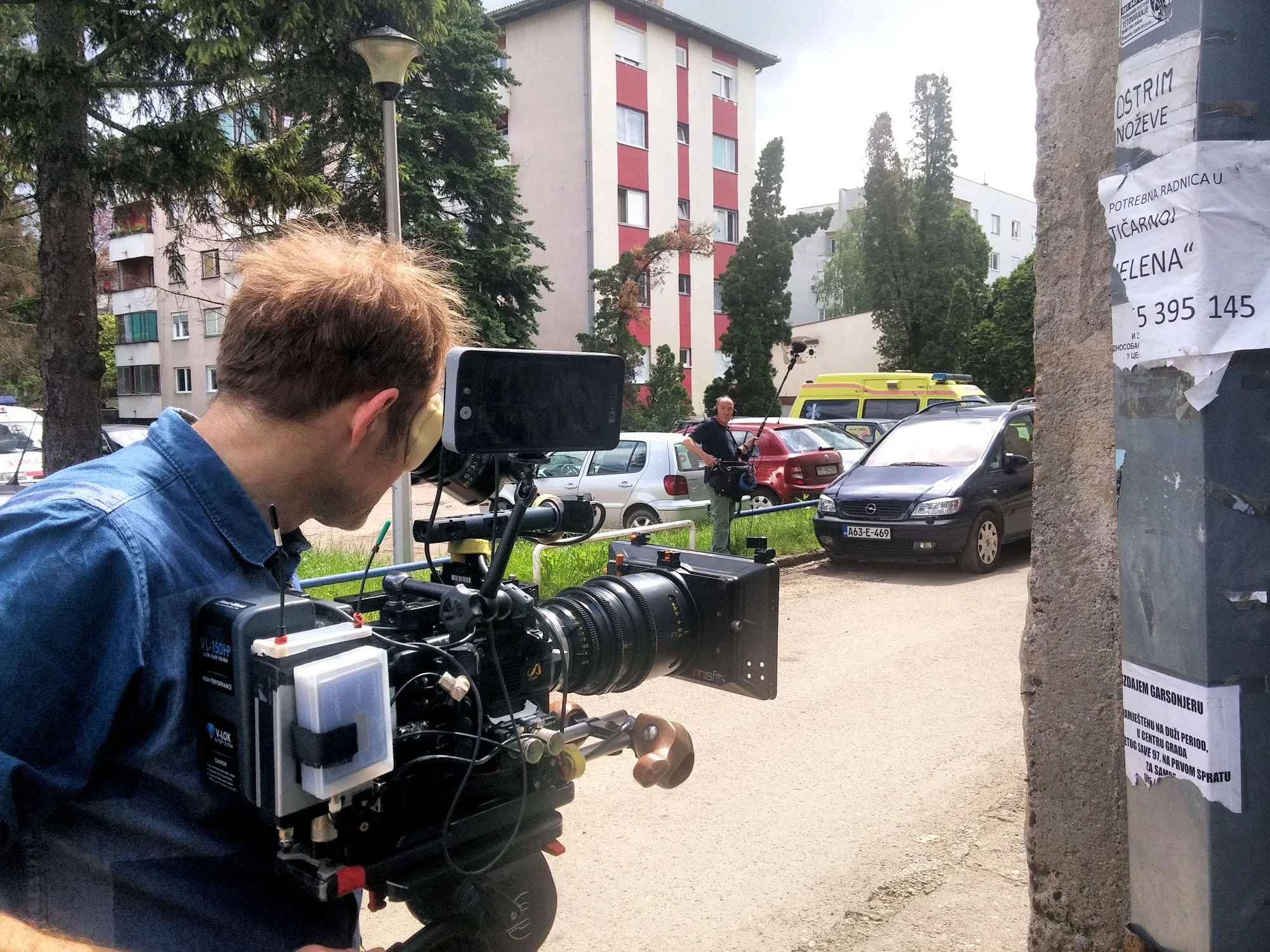 A man operating a professional video camera on a tripod outdoors in a residential area with parked cars, trees, and apartment buildings in the background. A person in the distance is holding a boom microphone.